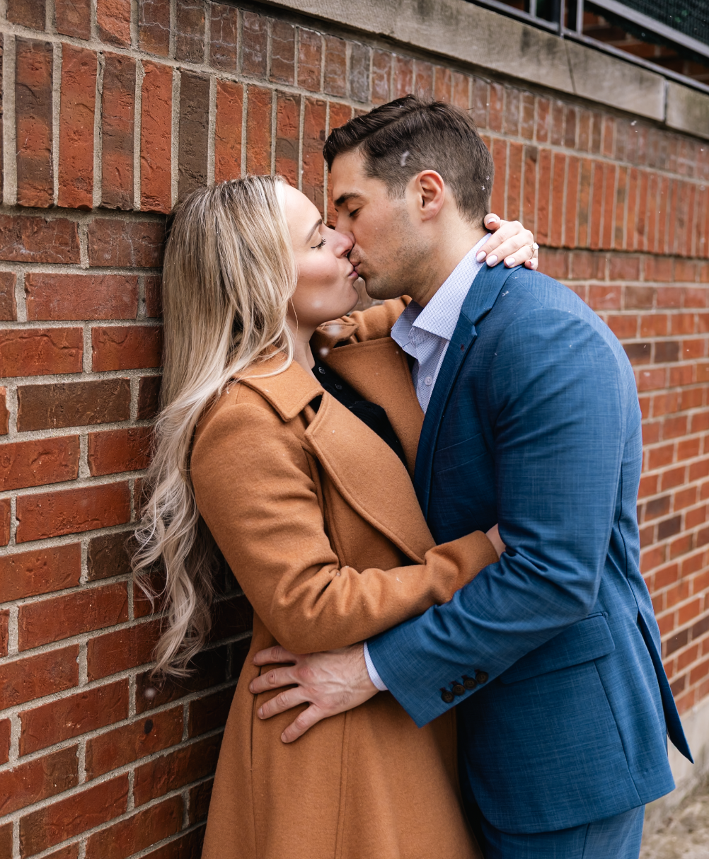 A couple kissing against a brick wall, the woman has long blonde hair and is wearing a brown coat, while the man is dressed in a blue suit.