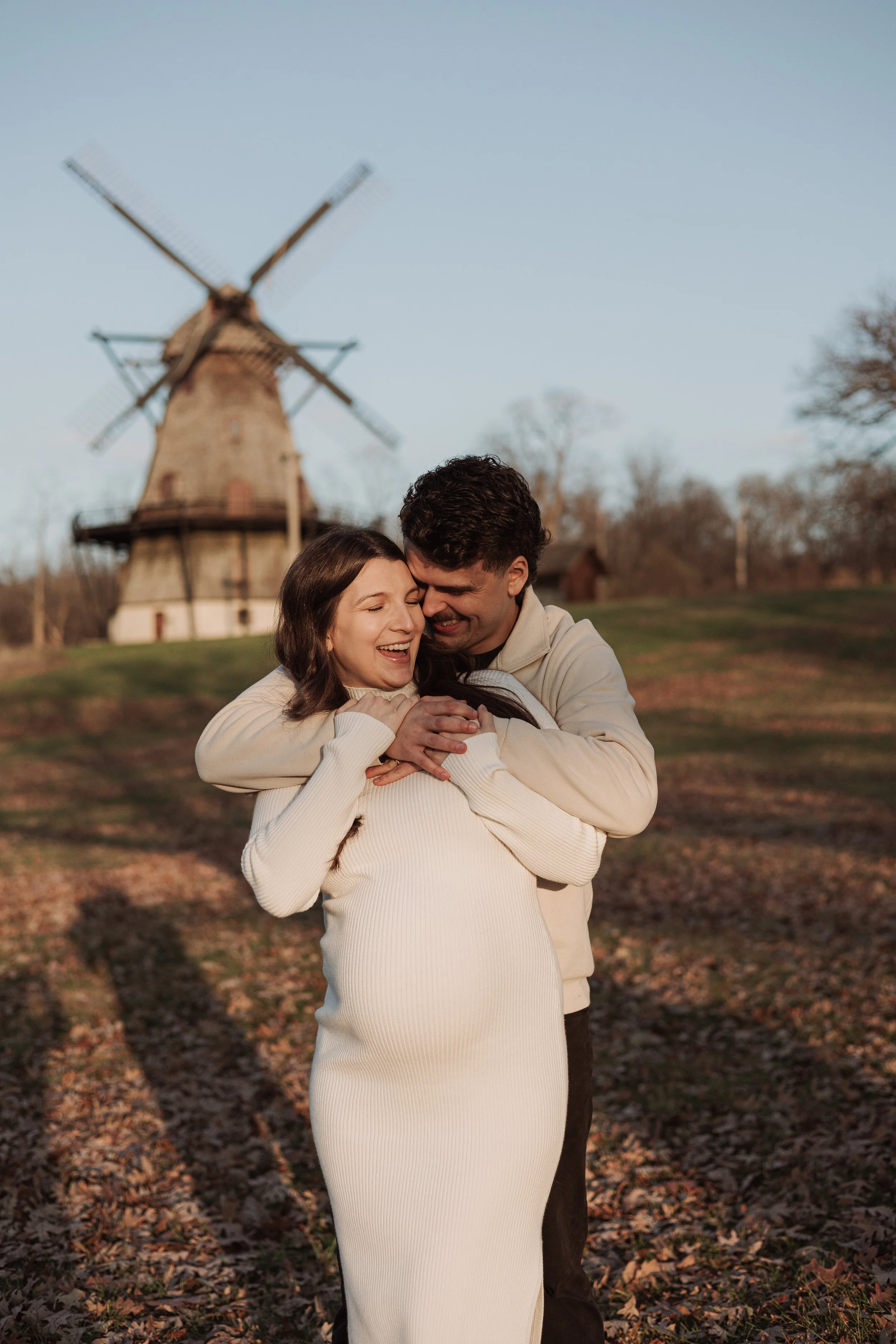 A happy couple embracing outdoors in front of a windmill, with trees and a clear sky in the background, during fall.