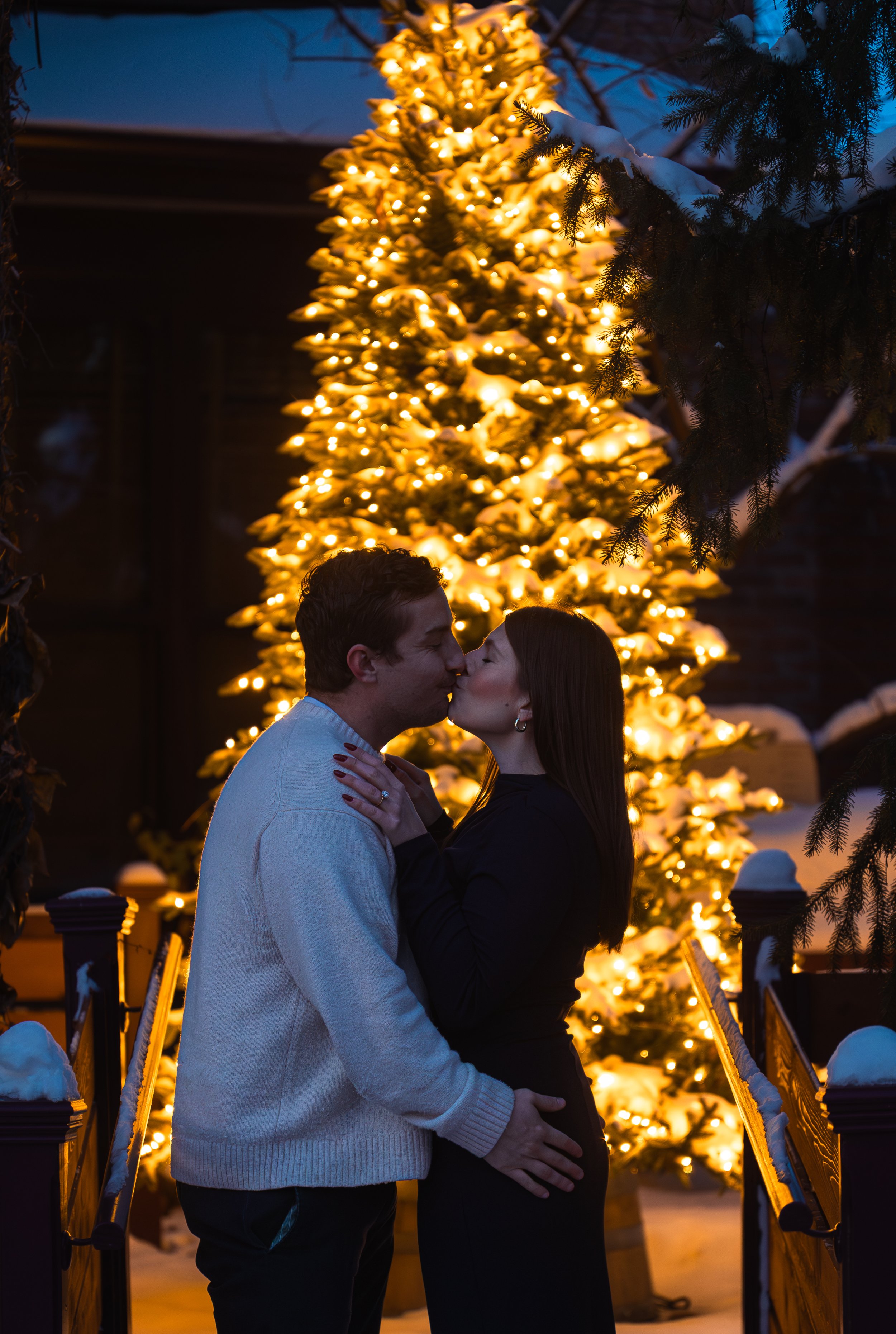 A couple kissing in front of a lit Christmas tree at night, with snow on the ground and trees in the background.