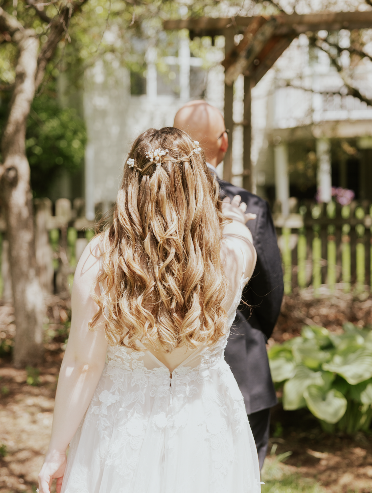 A bride and groom standing outdoors, with the bride adjusting the groom's tuxedo, featuring greenery and a wooden fence in the background.
