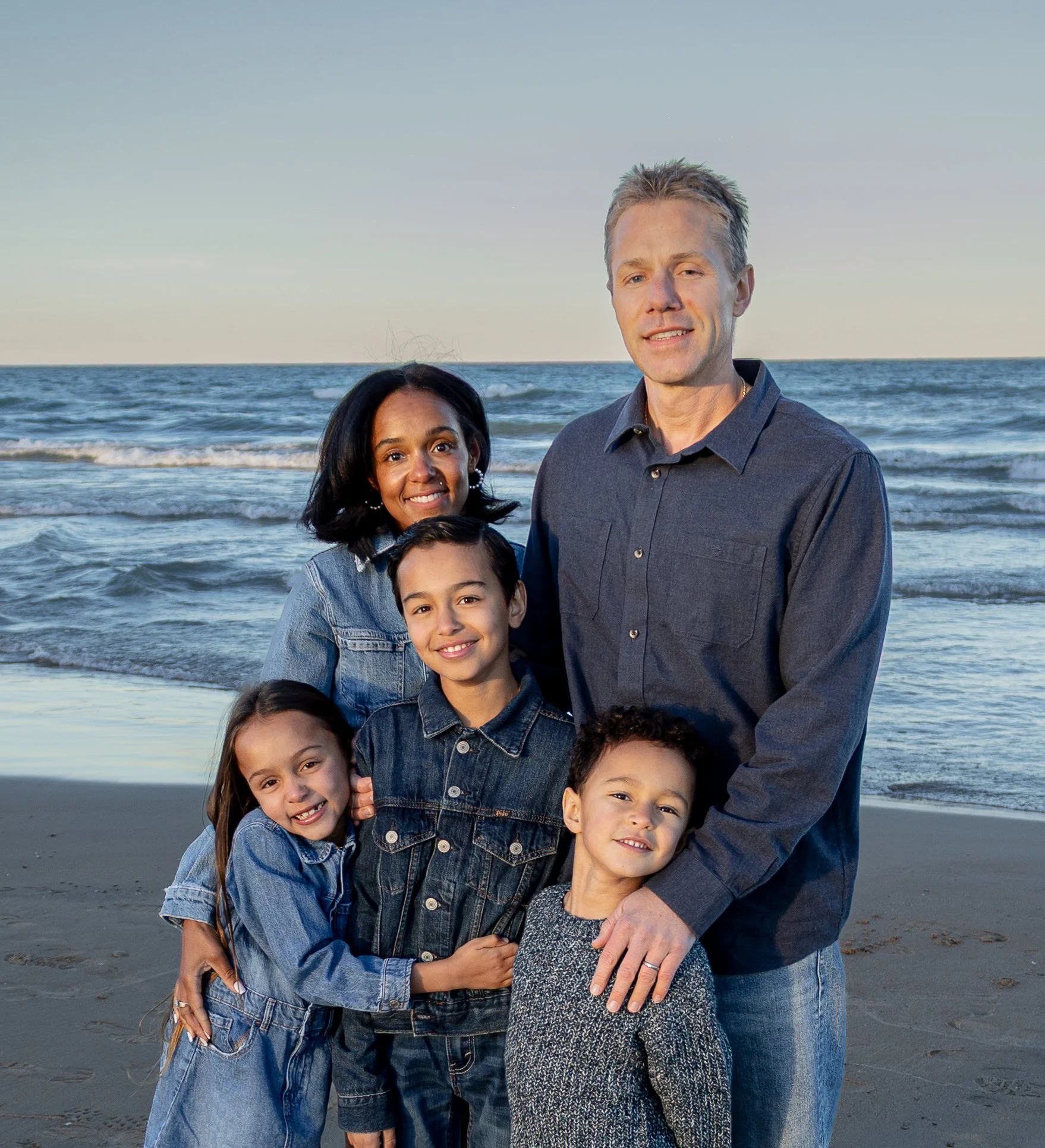 A family enjoying a day at the beach with children and adults smiling, standing close together, with the ocean in the background during sunset.