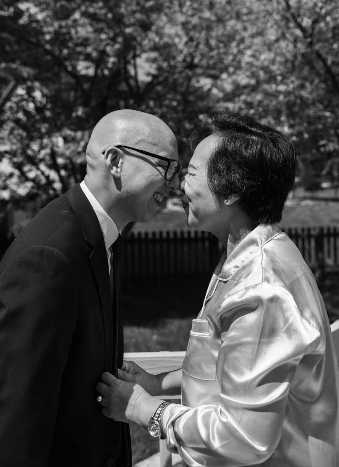 A man and woman face each other with foreheads touching, smiling in a park with trees and a fence in the background.