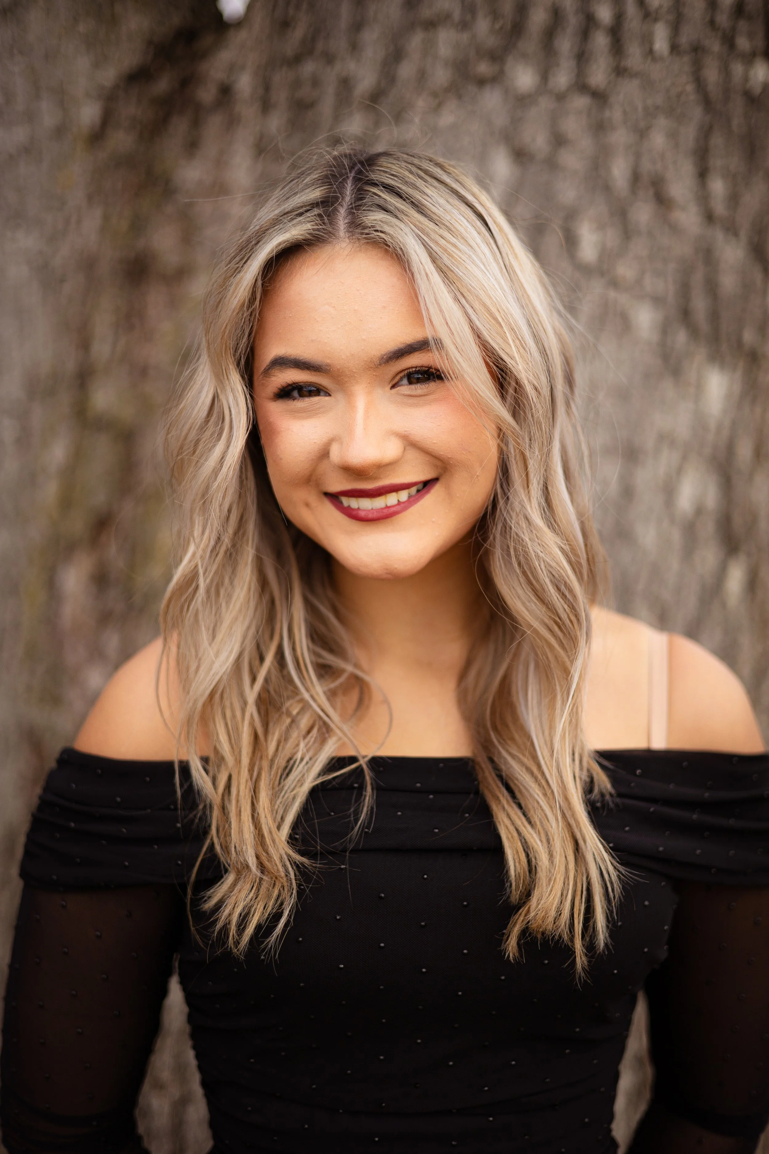 A woman with long blonde hair, wearing a black off-the-shoulder top with small embellishments, is smiling and standing against a background of tree bark.