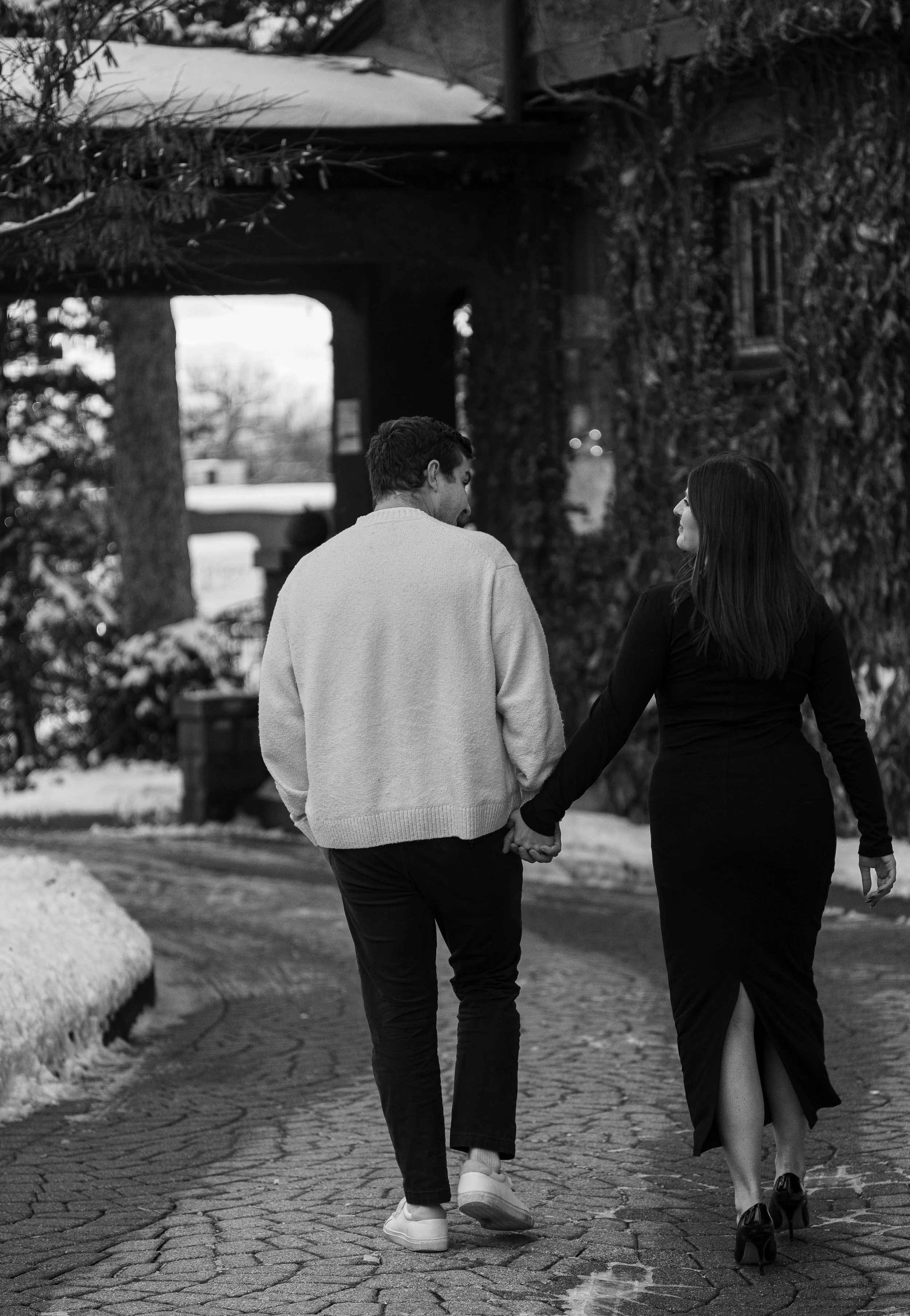 A couple walking hand in hand on a cobblestone path in winter, dressed stylishly, with leafless trees and a rustic wooden structure in the background.