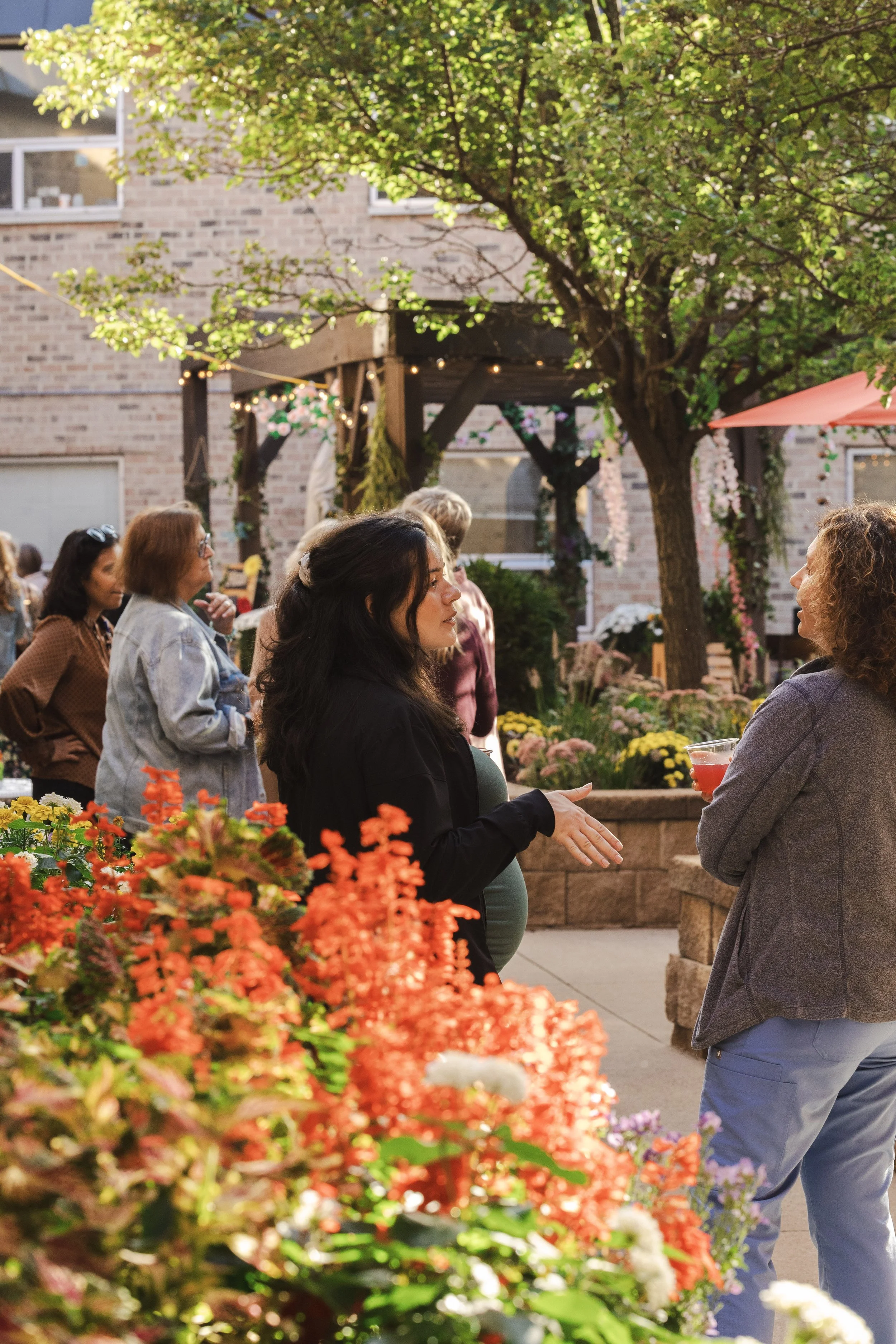 People enjoying conversation at an outdoor gathering next to a garden with colorful flowers and a large tree, with houses in the background.