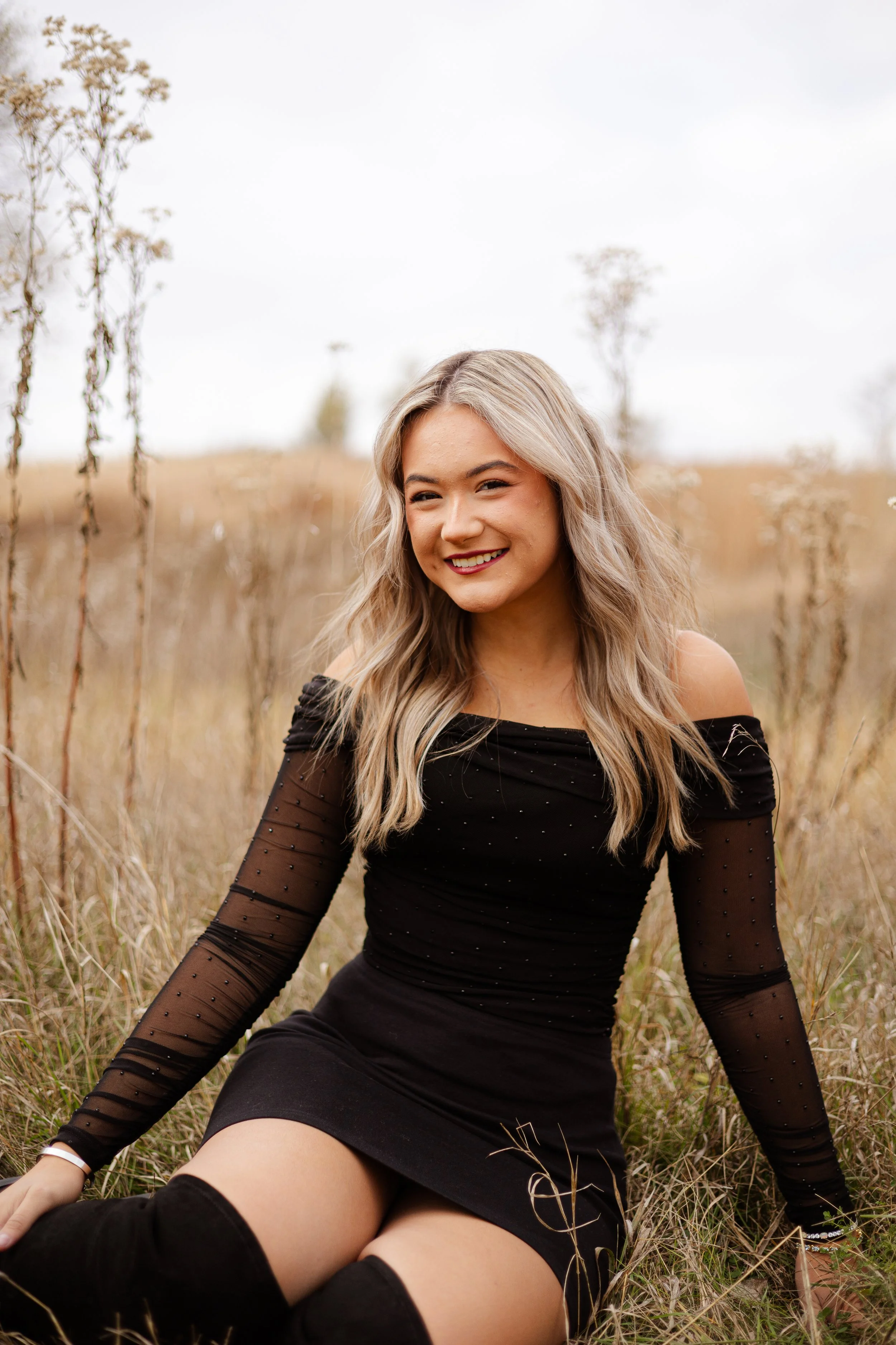 A smiling woman with long blonde wavy hair wearing a black dress with sheer sleeves, sitting outdoors in a field of tall grass and dried plants.