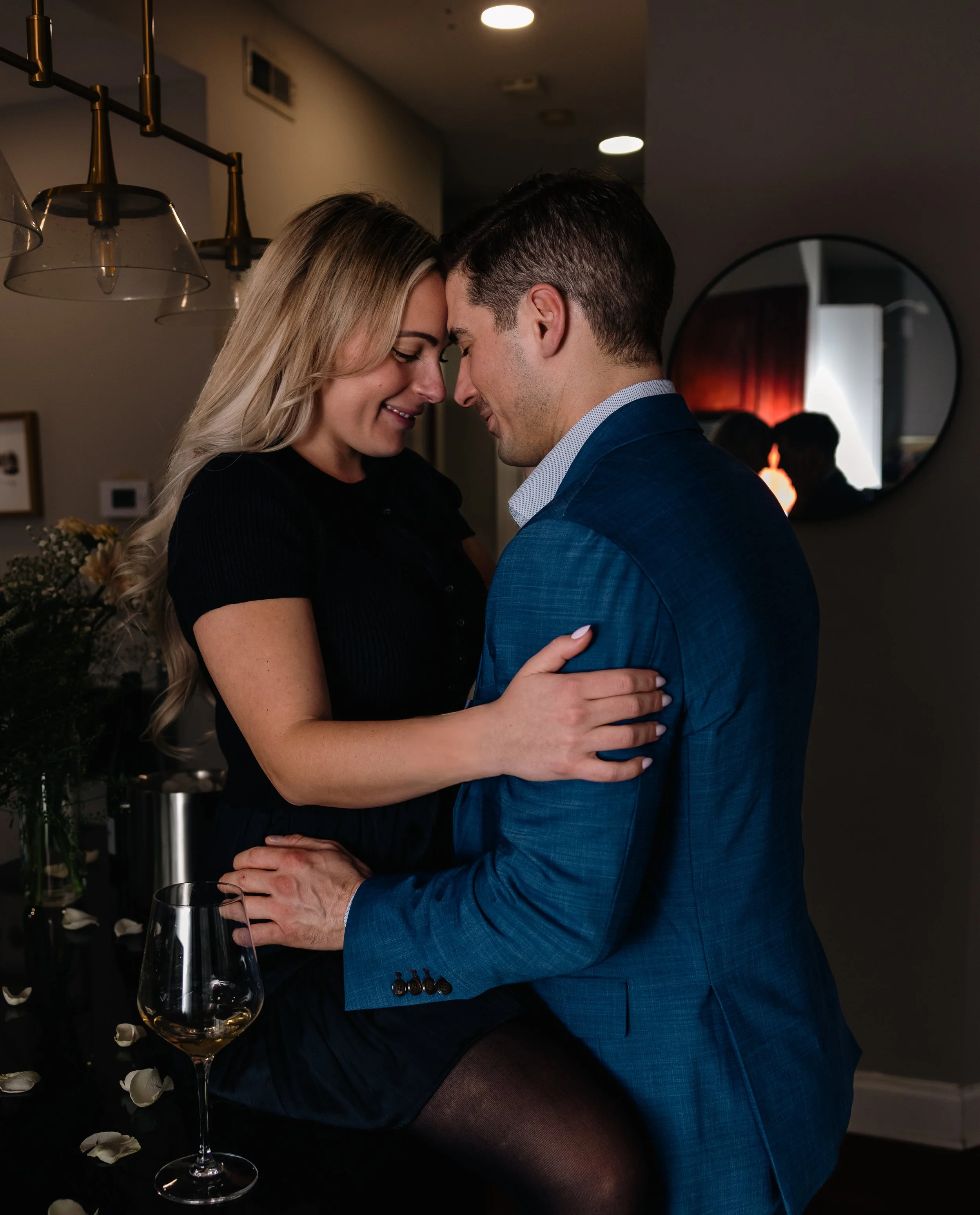A couple is intimate, with foreheads touching, in a romantic moment at a social gathering. The woman has long blonde hair and is wearing a black dress, and the man is dressed in a blue suit. They are close together, smiling and holding each other gen