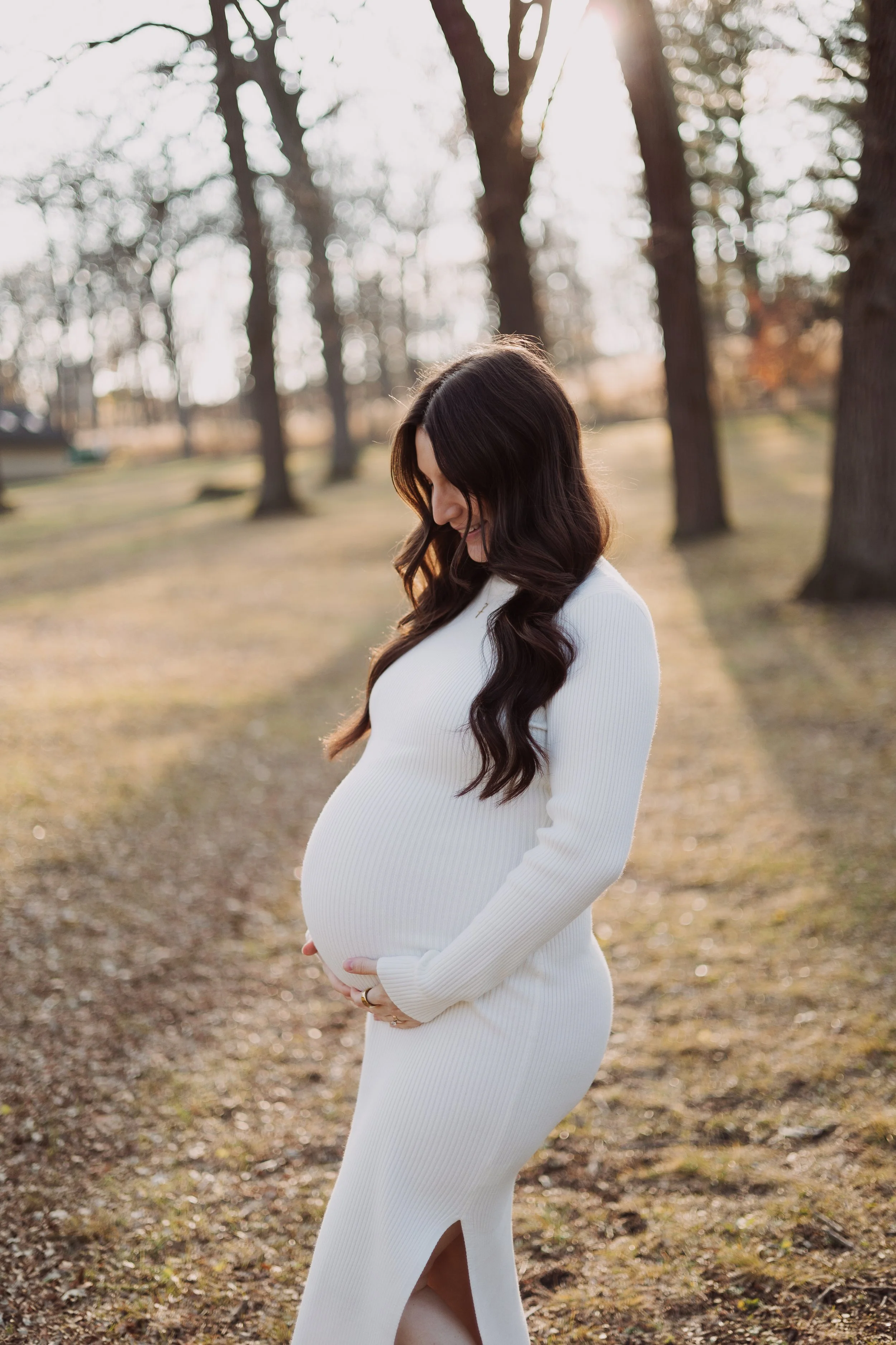 A pregnant woman with long wavy brown hair smiling and looking down while standing outdoors in a park with tall trees and sunlight filtering through