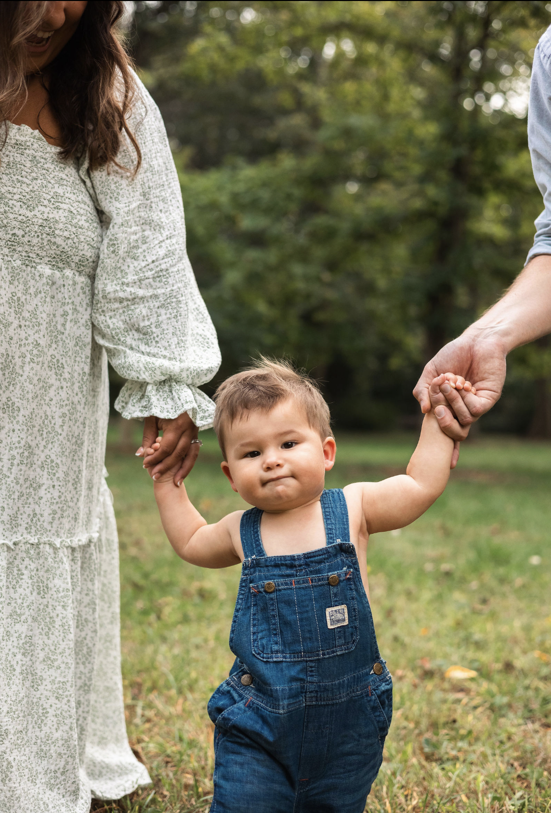 A young boy holding hands with two adults outdoors in a park, with trees in the background.
