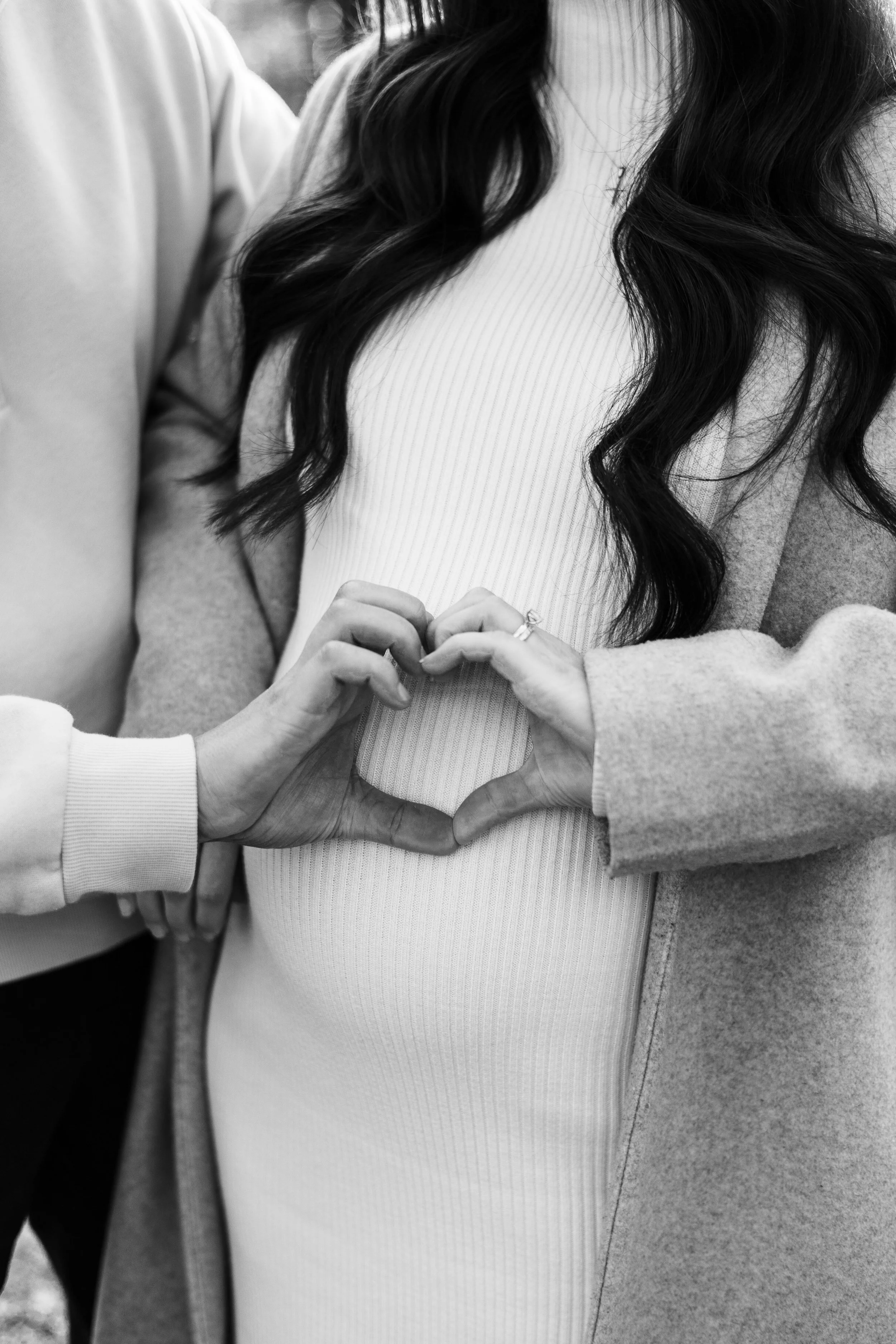 Close-up of two women forming a heart shape with their hands over the pregnant woman's belly, both wearing cozy clothing, black and white photo.
