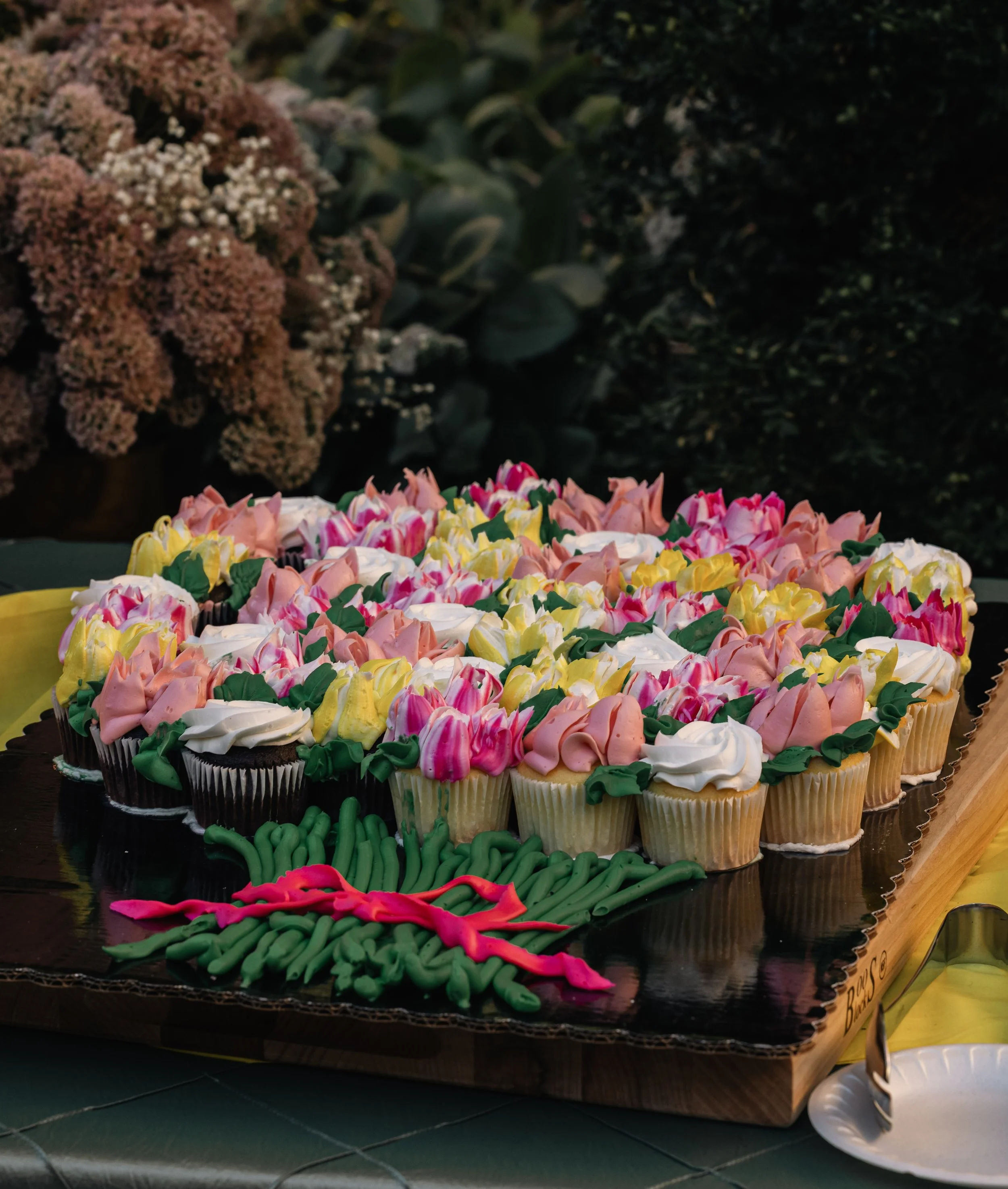 Colorful cupcakes topped with pink, white, yellow, green, and brown swirled frosting, arranged on a black tray with green and pink decorative icing. In the background, there are large pinkish flowers and dark green leafy plants. A serving spoon is vi