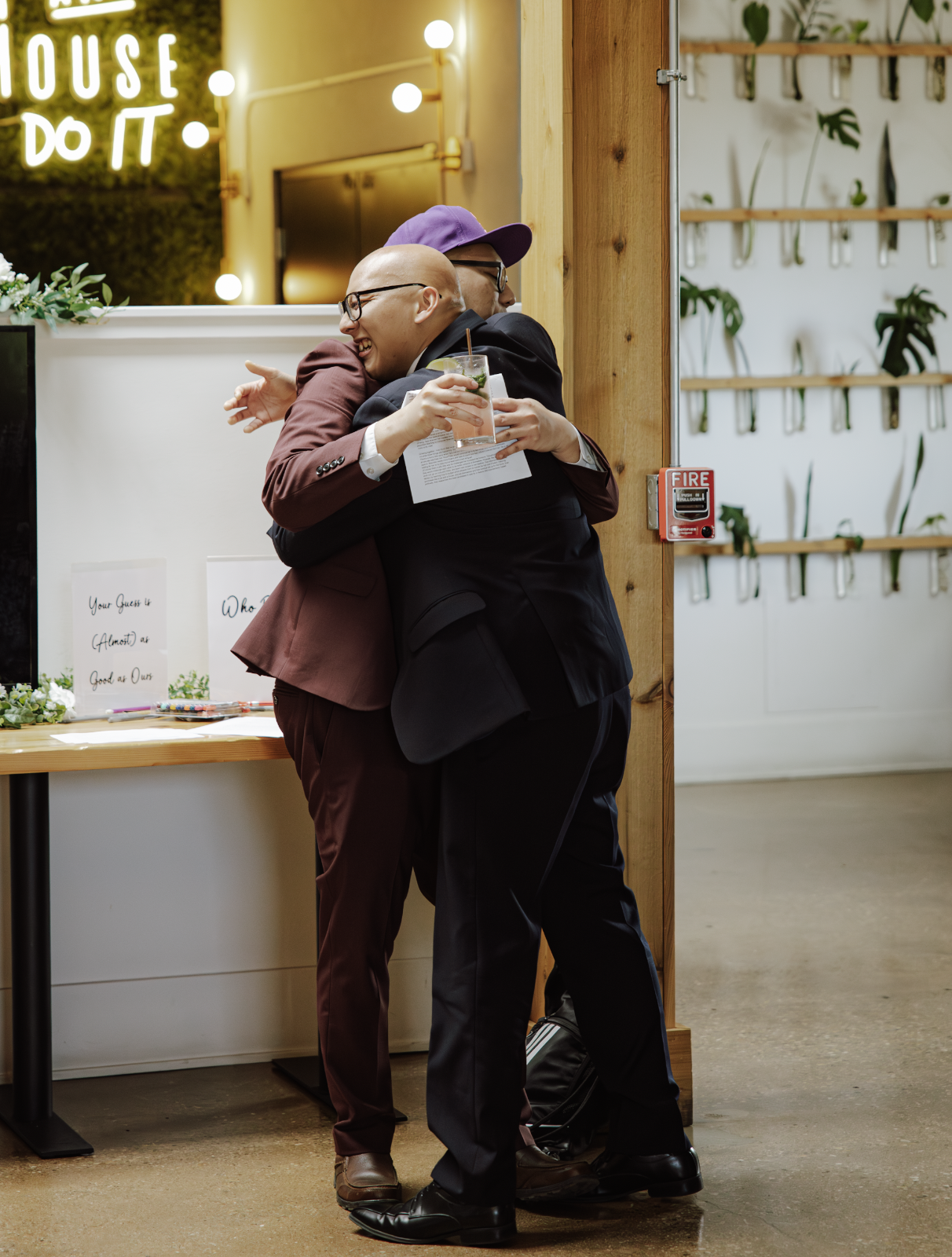 Two men in suits hugging and smiling in an indoor setting, one holding a beverage, near a wall with a plant display and a sign that reads 'HOUSE DO IT' in bright lights.