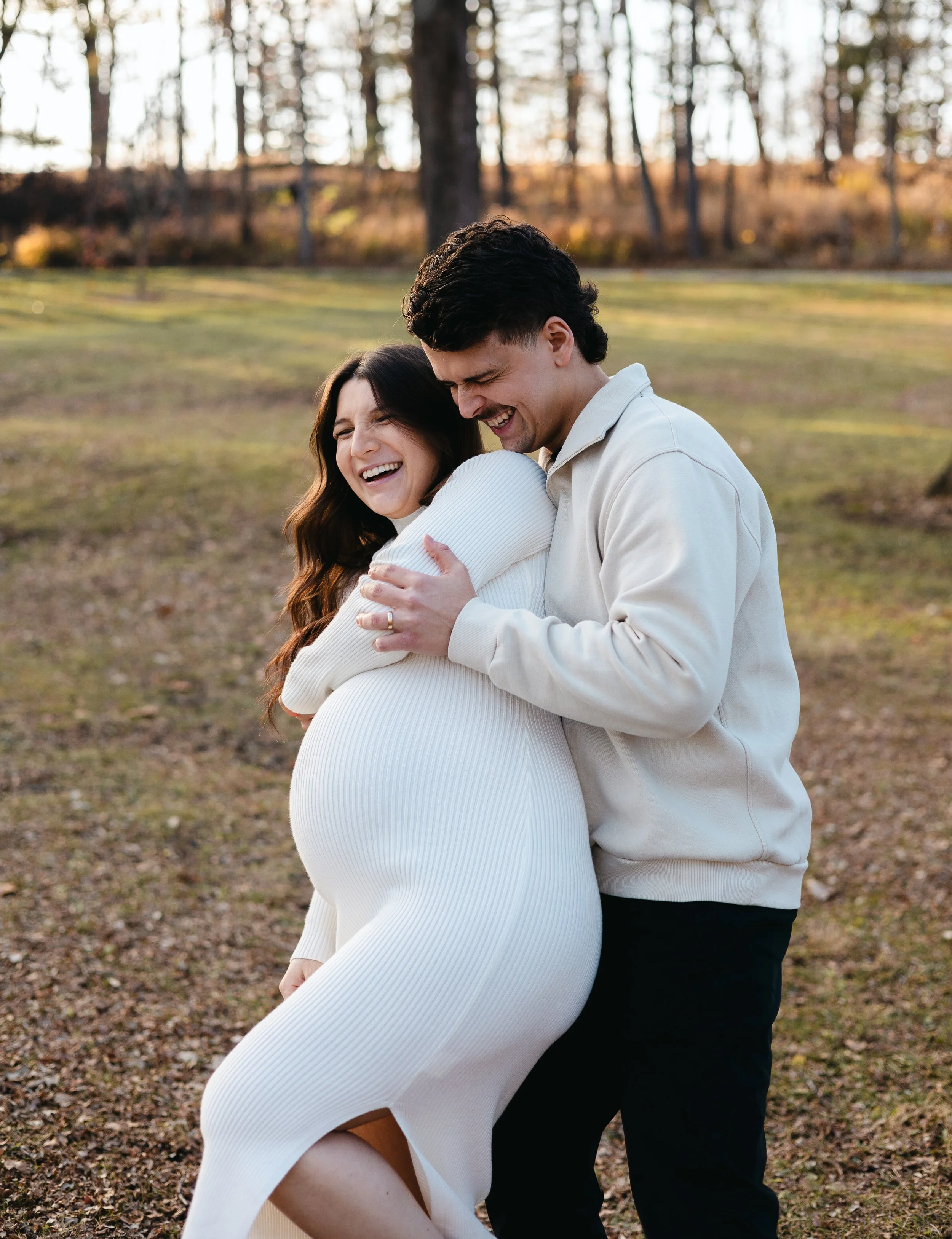 A couple sharing a joyful hug outdoors in a wooded park during autumn, with trees and fallen leaves in the background.
