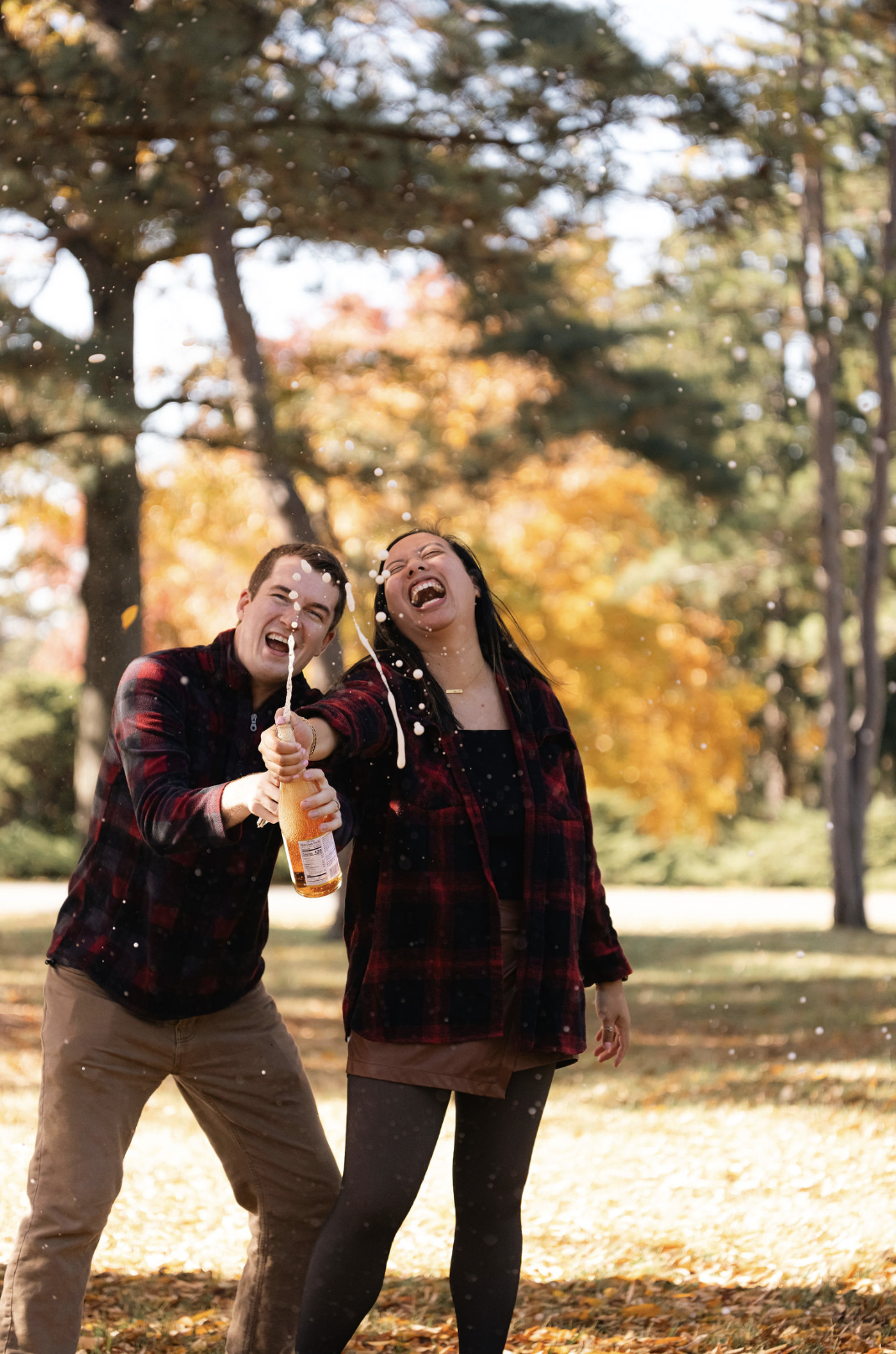 Two people celebrating outdoors in fall, spraying champagne, with colorful autumn leaves and trees in the background.