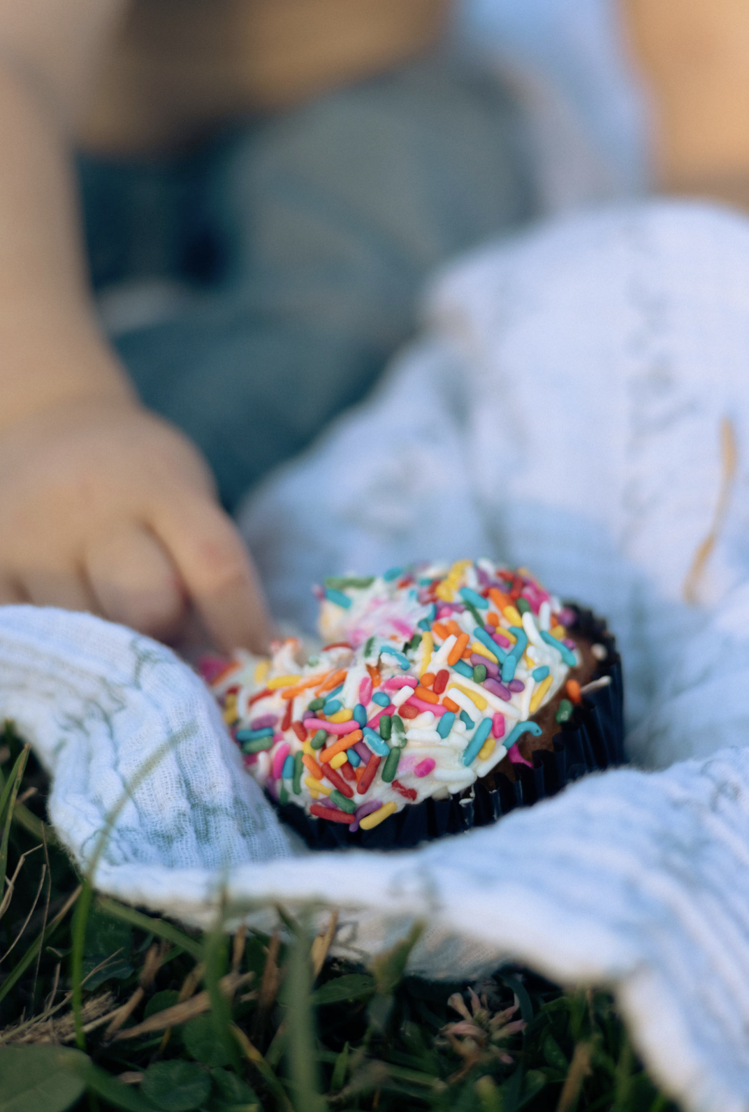Close-up of a child's hands holding a cupcake with white frosting and colorful sprinkles, lying on a white cloth outdoors on grass.