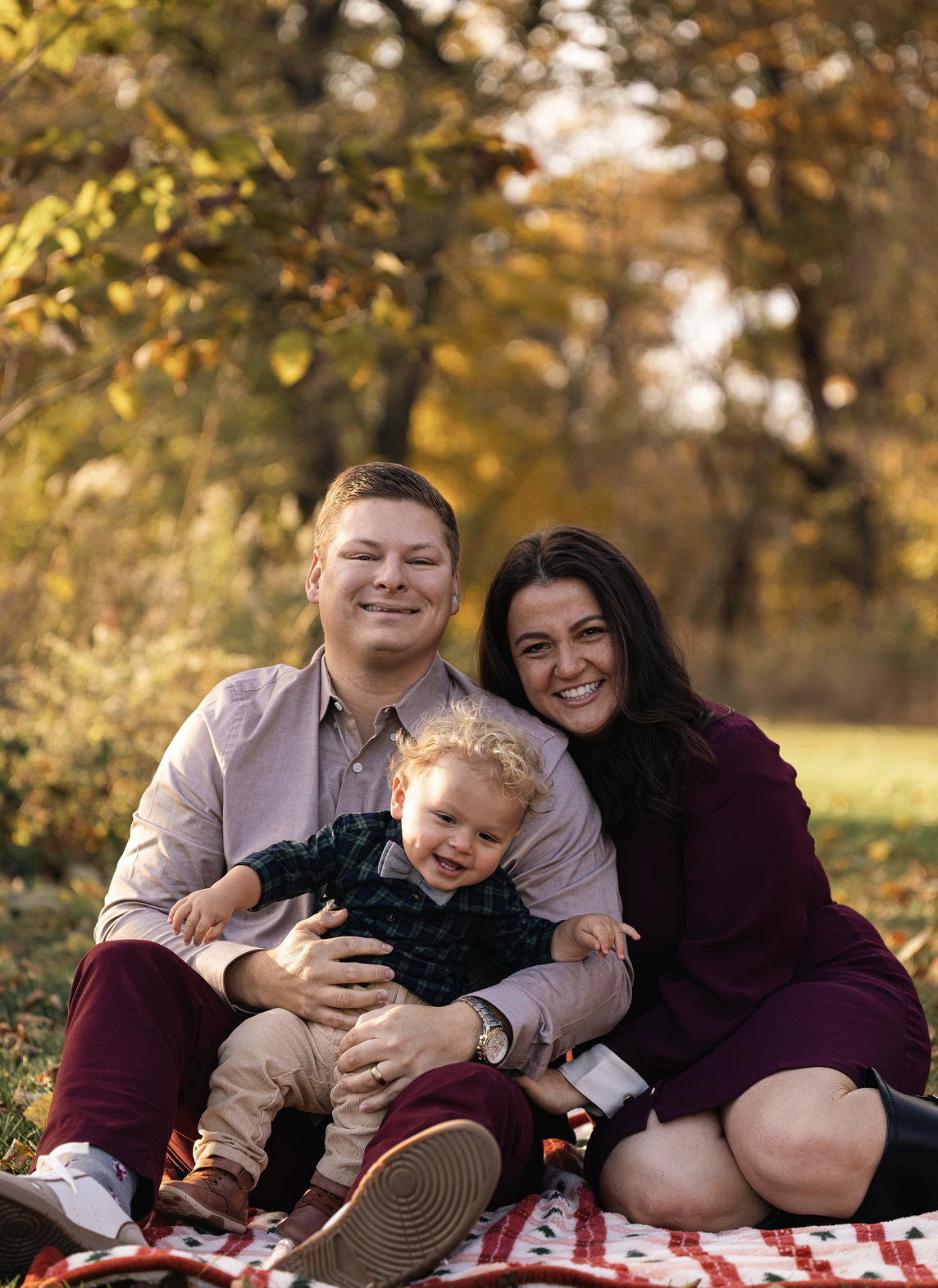 Family of three sitting on a red and green plaid blanket outdoors in autumn, smiling. The man is on the left, the woman on the right, and a young boy with curly blonde hair and a dark checkered shirt in the middle. Trees with golden leaves are in the