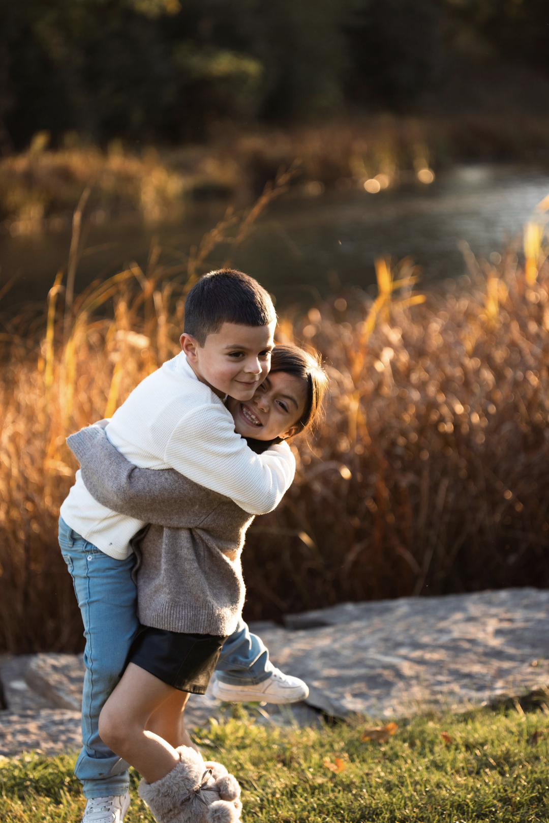 Two children, a boy and a girl, playing by a river at sunset. The boy is giving the girl a piggyback ride and they are smiling happily.