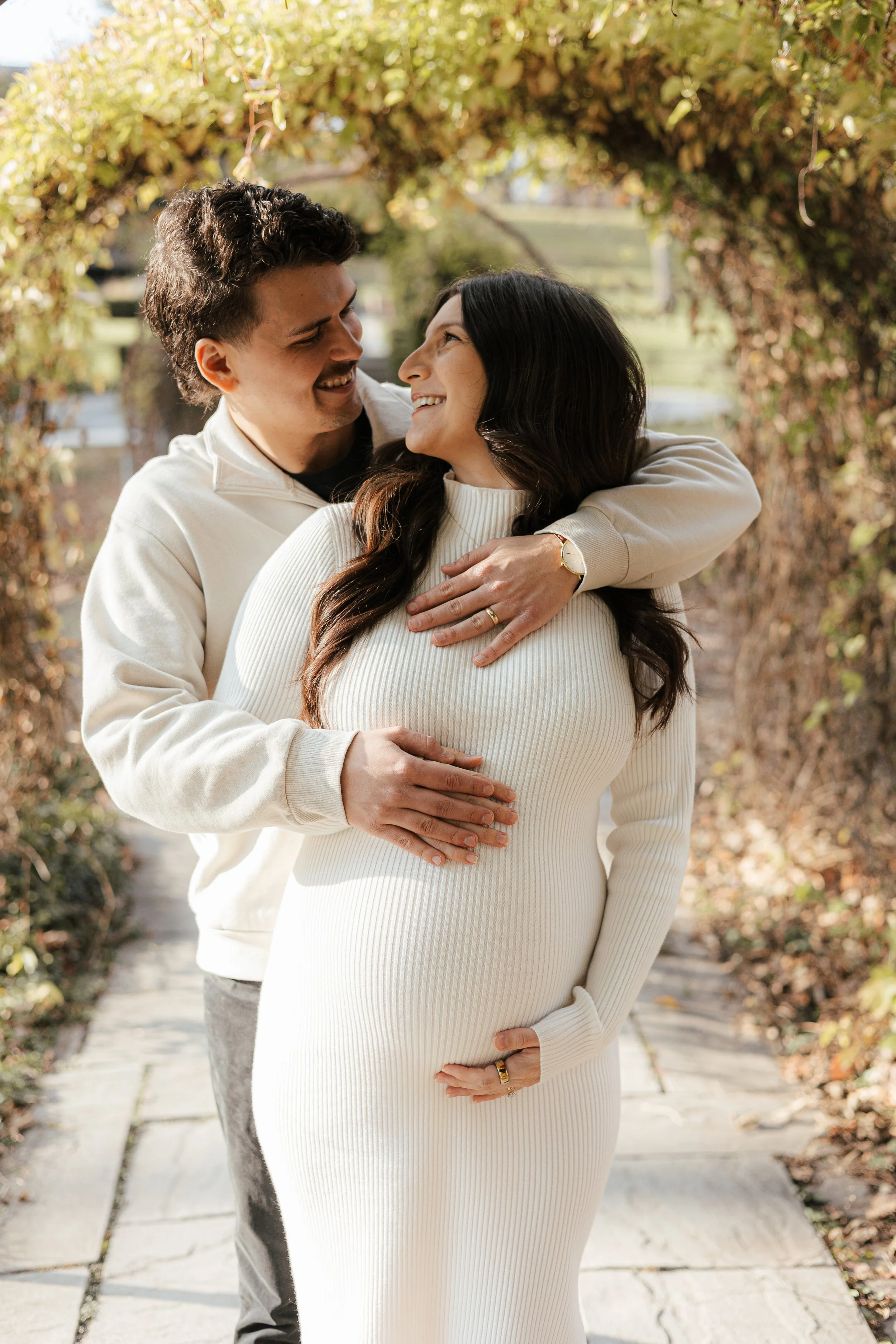 A couple stands close together outdoors on a sunny day, with the woman visibly pregnant. The man has his arms around her, and they are smiling at each other. The background features a pathway and an arch made of leaves with autumn-colored foliage.