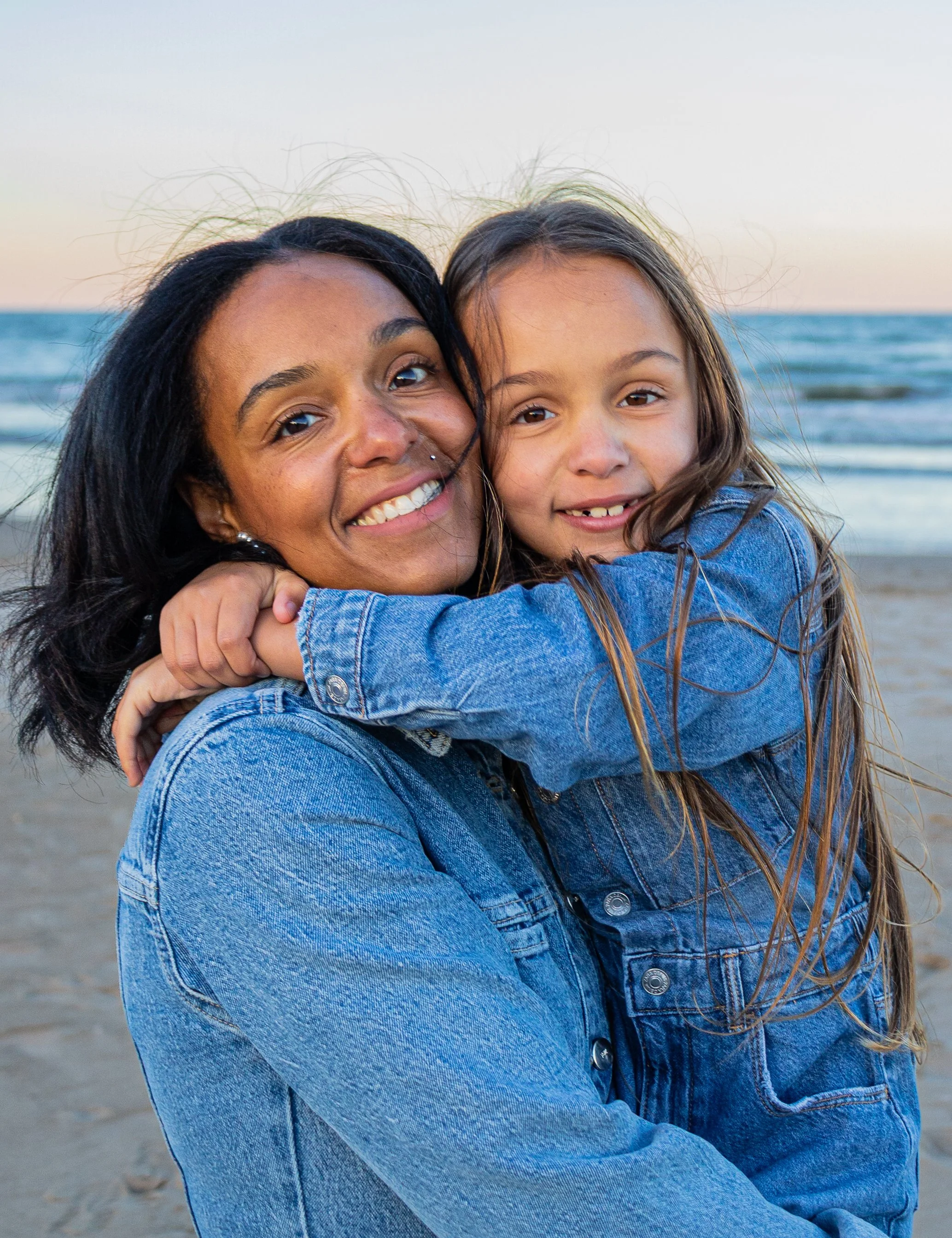 A woman and a young girl hugging on the beach with the ocean in the background during sunset or sunrise.