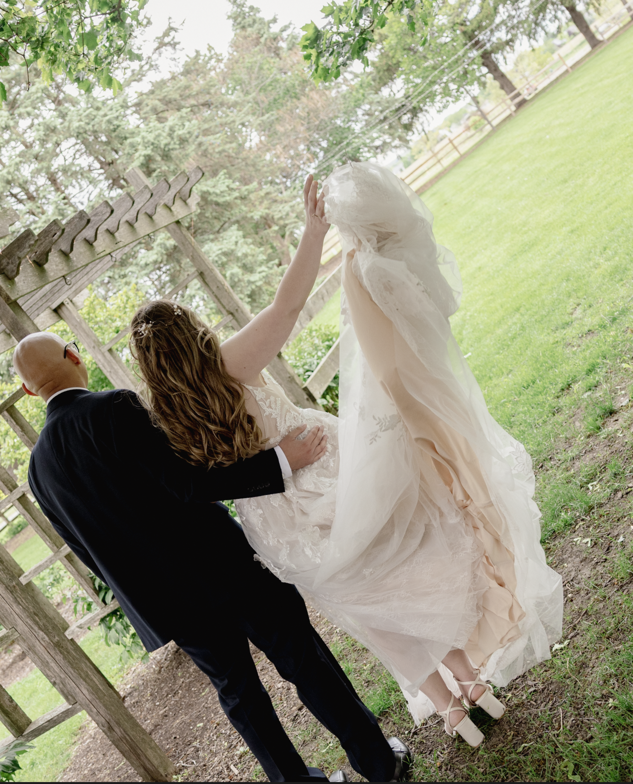 A newlywed couple dancing outdoors under green trees with a wooden trellis in the background. The bride wears a white wedding gown and high heels, while the groom wears a dark suit and glasses. The bride has brown hair and is lifting her hand towards