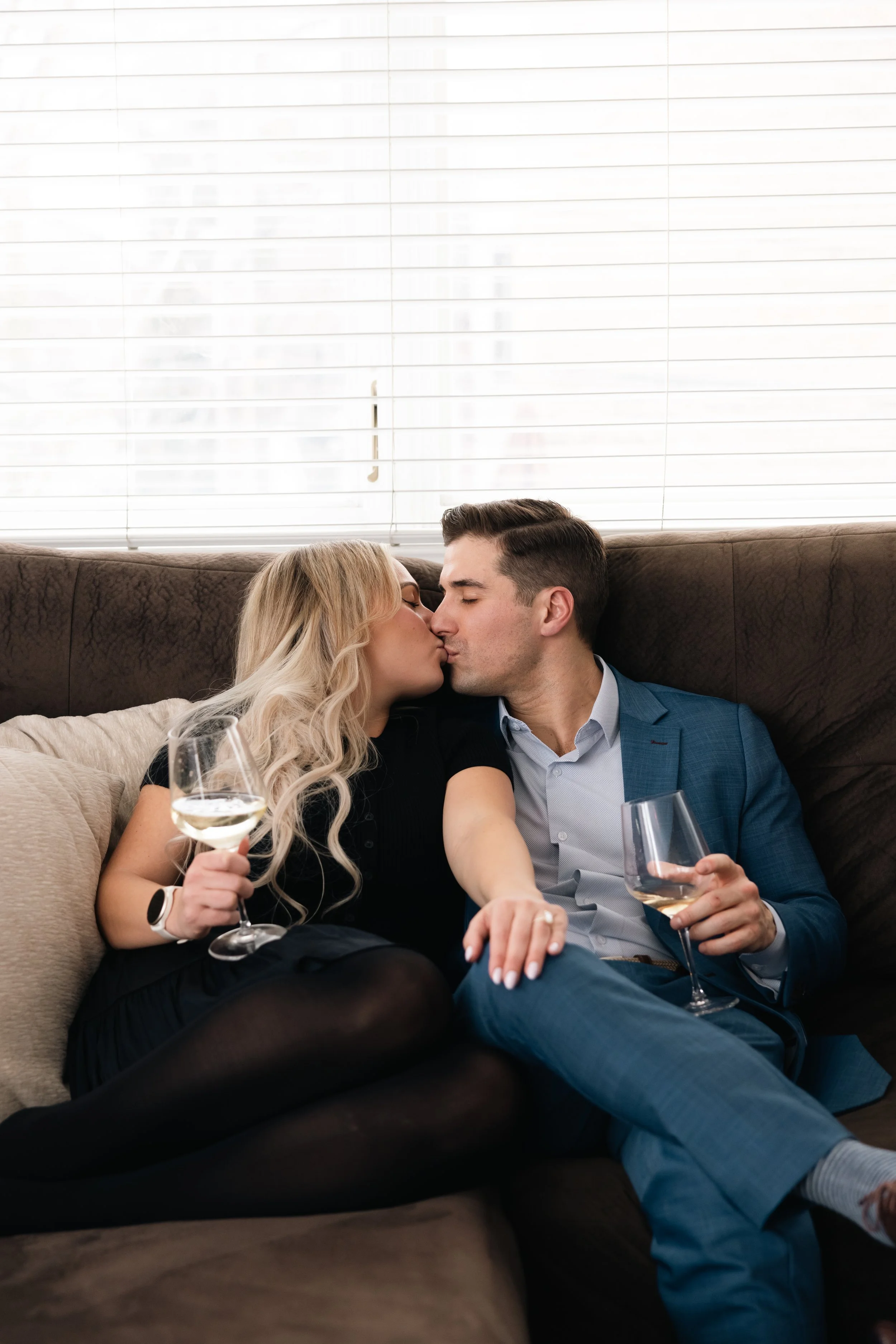 Couple sharing a kiss on a sofa with wine glasses in hand, against a background of white window blinds.