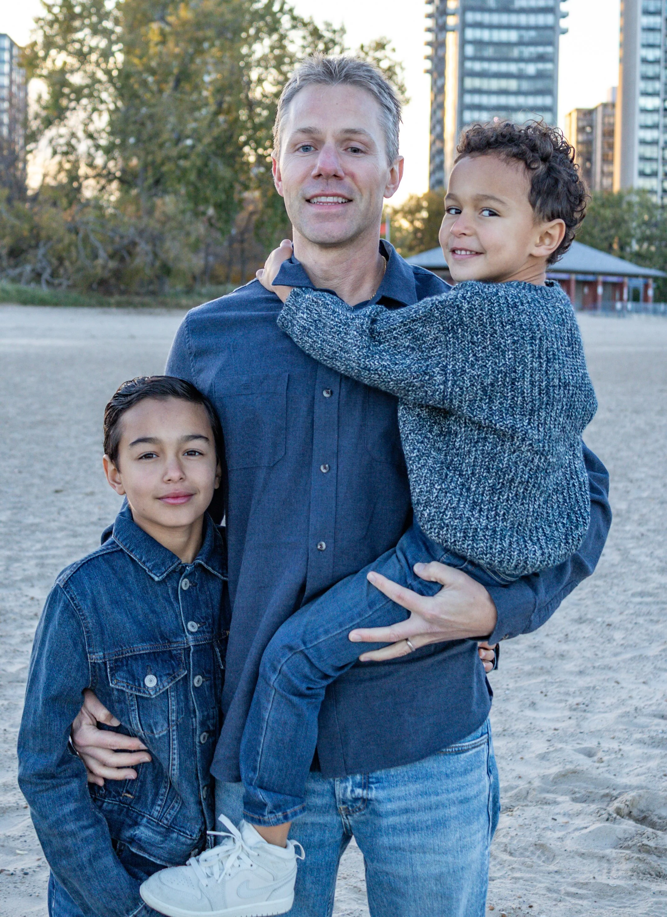 A smiling man with two young boys on a sandy outdoor area, with trees and tall buildings in the background during daylight.