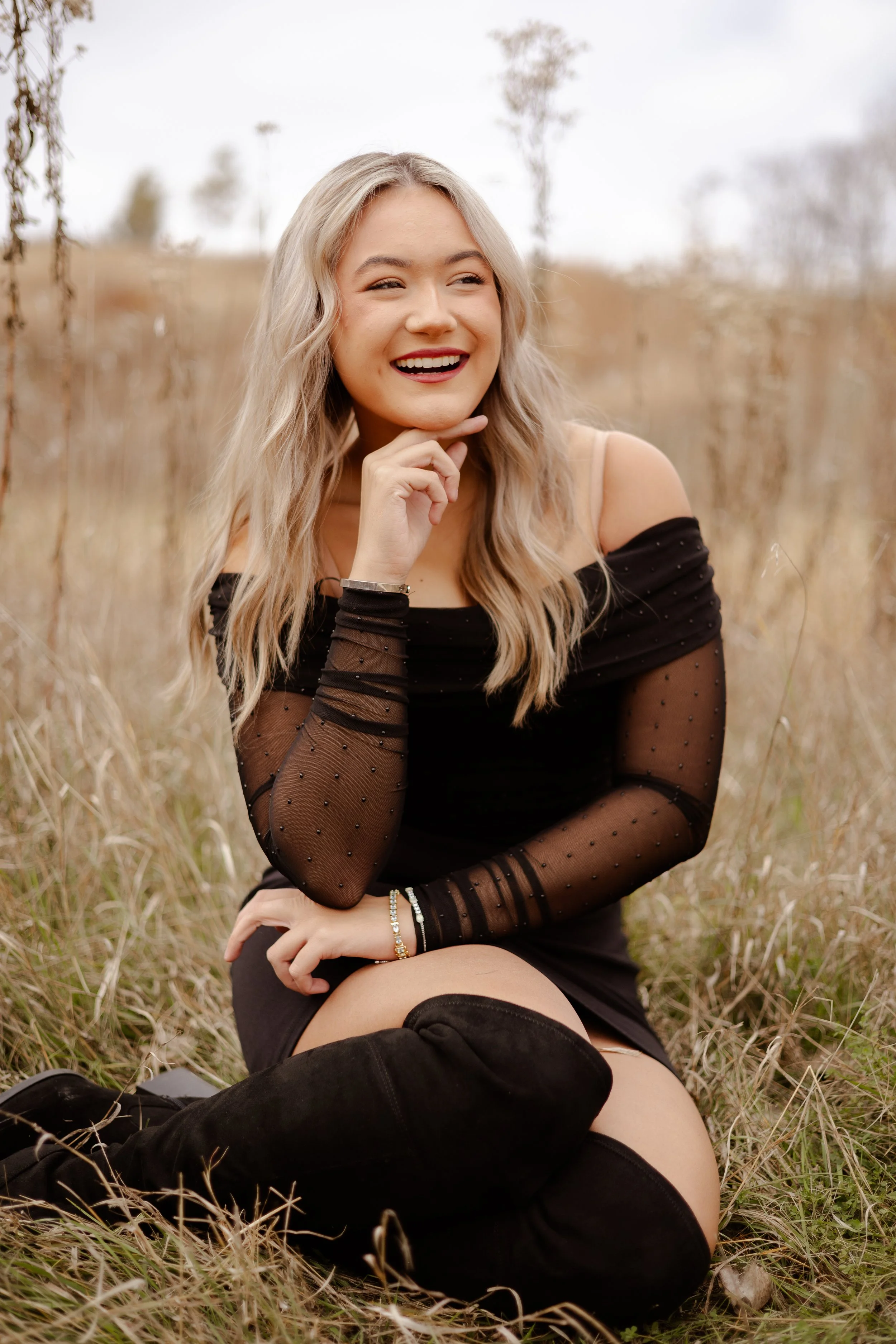 A young woman with blonde hair, wearing a black off-shoulder top with sheer polka-dot sleeves, sitting in a field of tall, dry grass. She is smiling and looking to the side, with her chin resting on her hand.