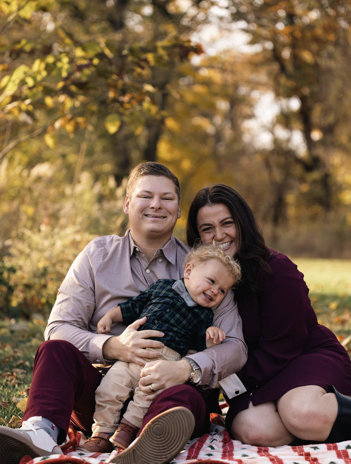Family of three sitting on a blanket outdoors in a park during fall, smiling at the camera. The father has short hair and is wearing a light shirt, the mother has long dark hair and is wearing a dark dress, and their young son with curly blonde hair 