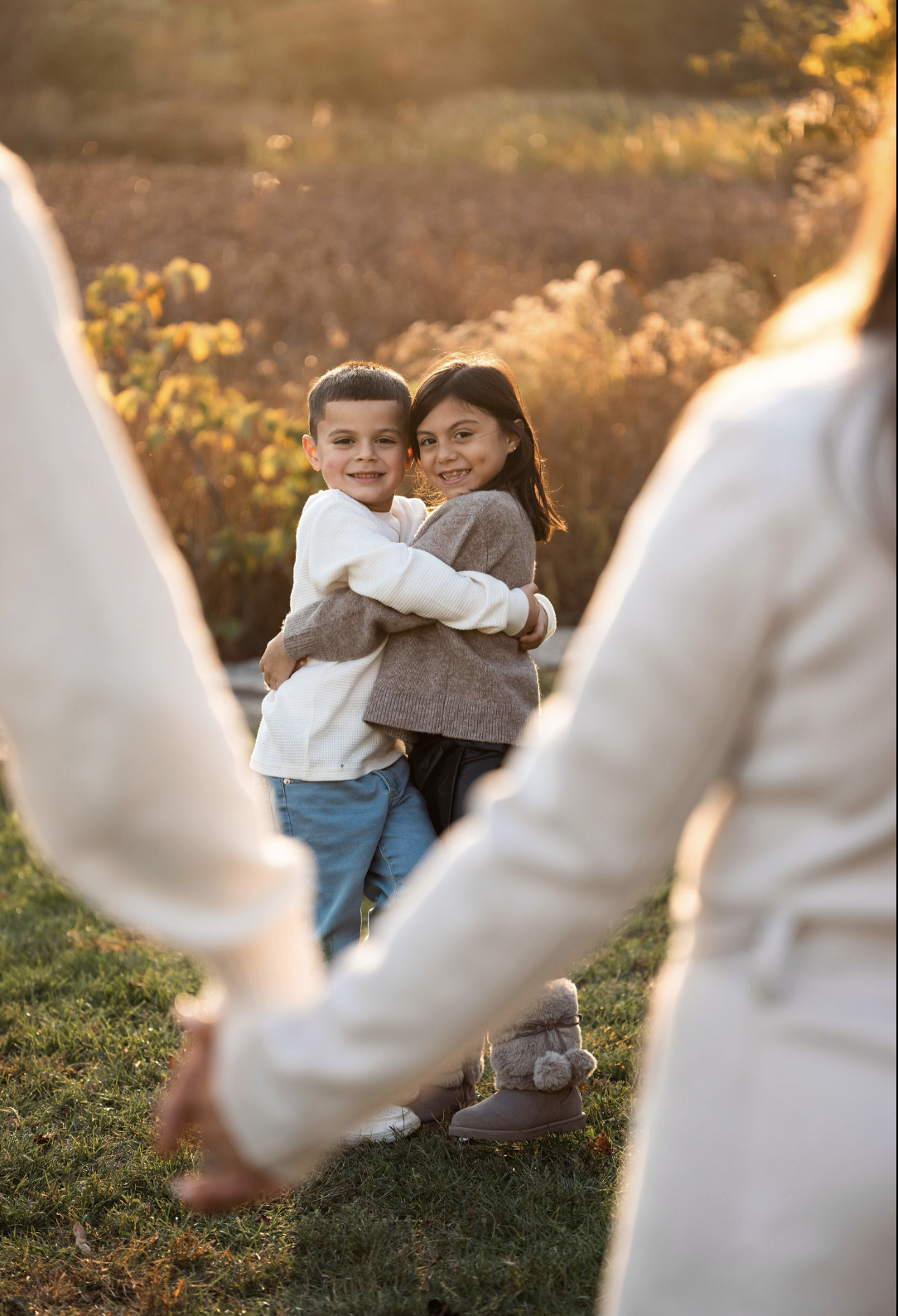 Two children, a boy and a girl, hugging each other and smiling at the camera, framed by adults holding hands in the foreground, with an outdoor background and warm sunset lighting.