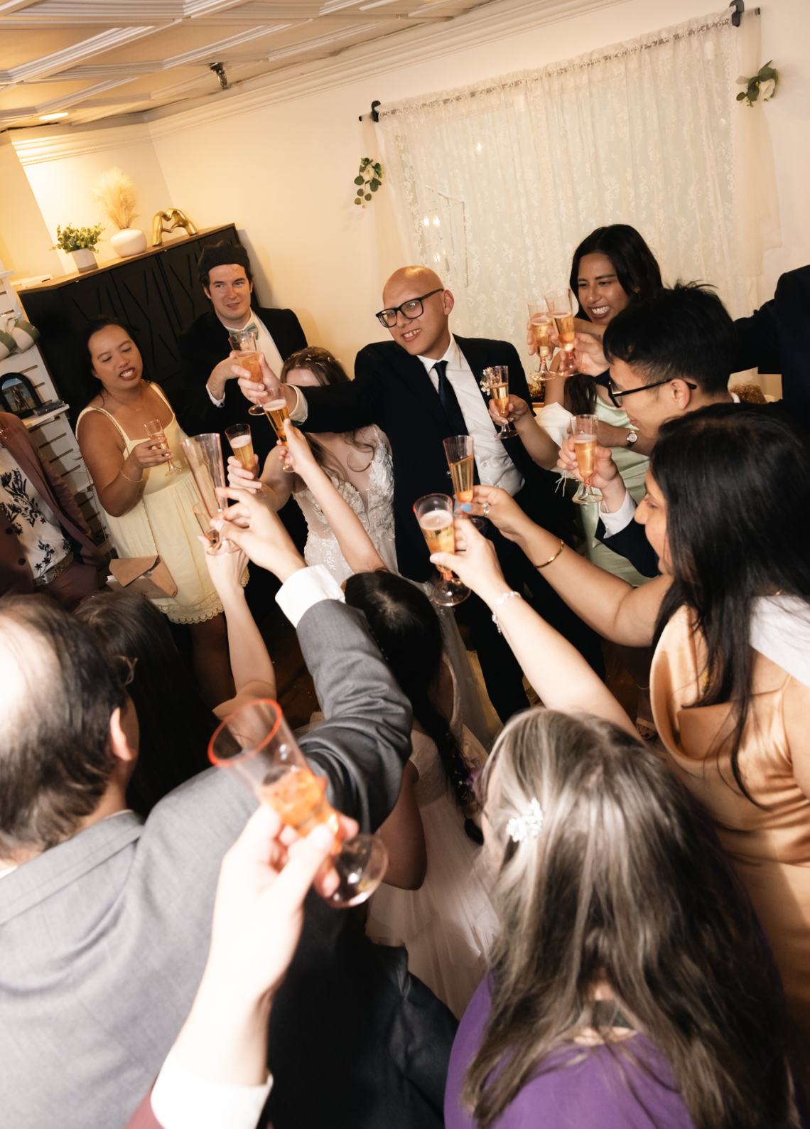 People celebrating at a party, raising glasses of champagne in a toast, in an indoor setting with warm lighting and curtains.