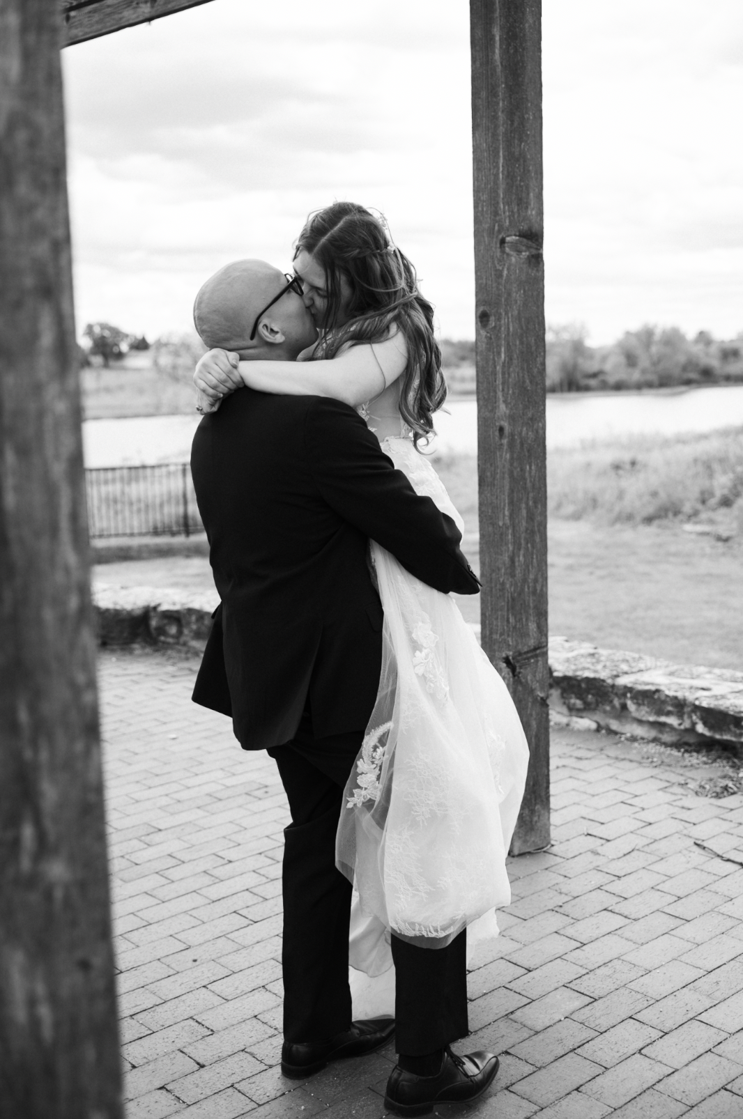A black and white photo of a man in a tuxedo lifting and kissing a woman in a wedding dress outdoors near water, framed by wooden posts.
