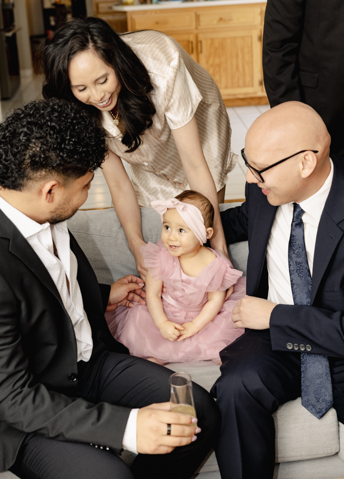 A group of adults and a baby girl are gathered together chatting indoors, with the baby dressed in a pink dress and a headband.