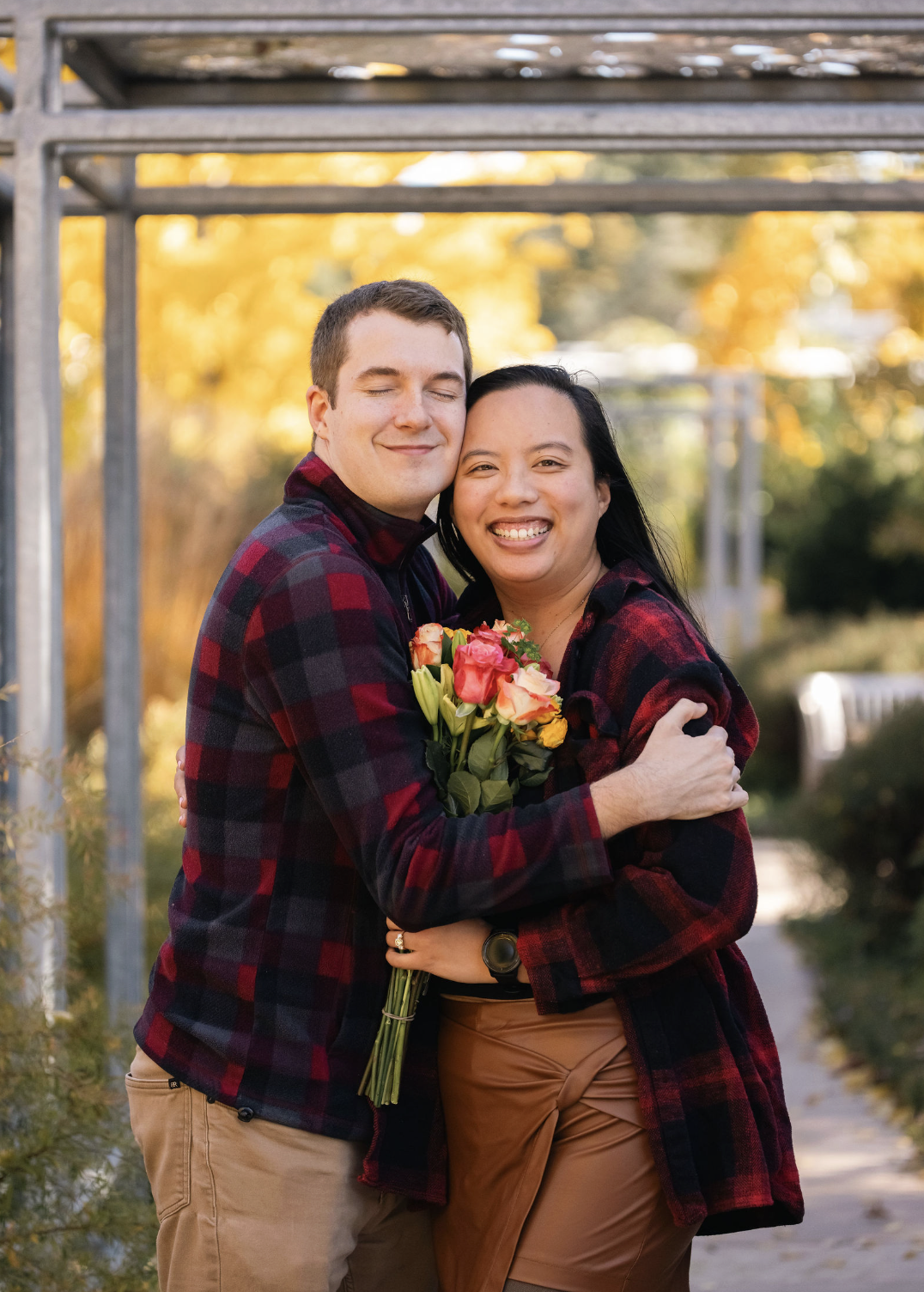 A couple hugging outdoors during fall, holding a bouquet of colorful flowers, both wearing matching plaid jackets, smiling with trees and a walkway in the background.