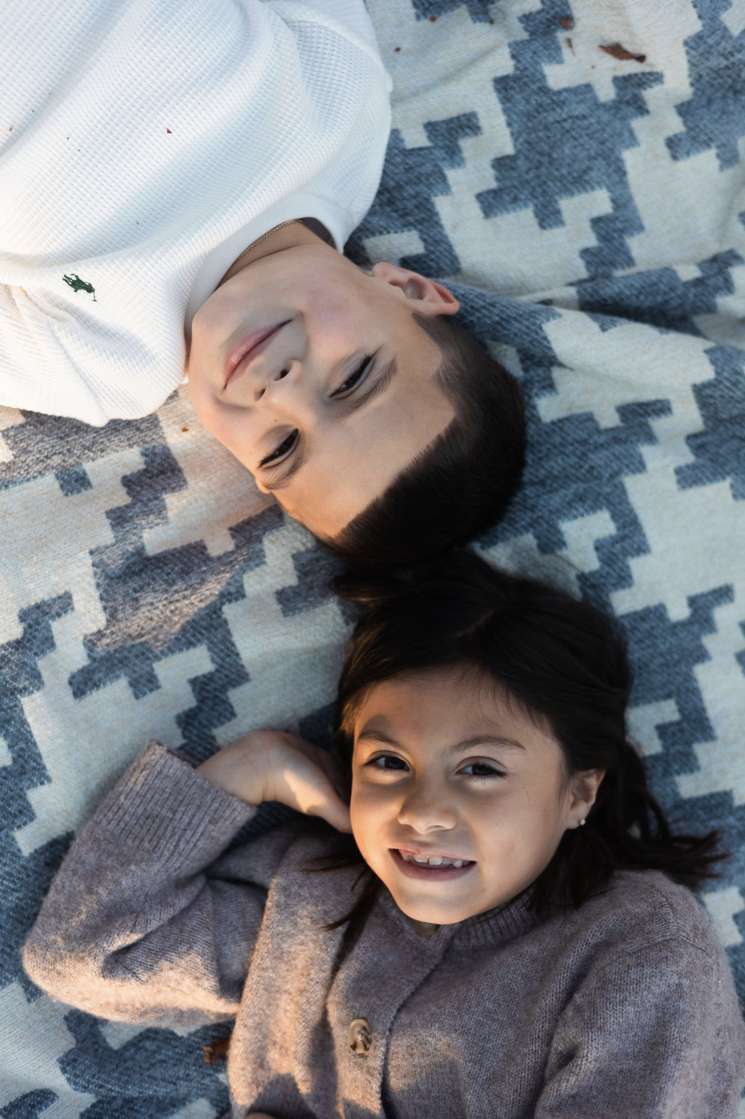 A boy and girl lying on a patterned blanket, looking up at the camera, smiling.