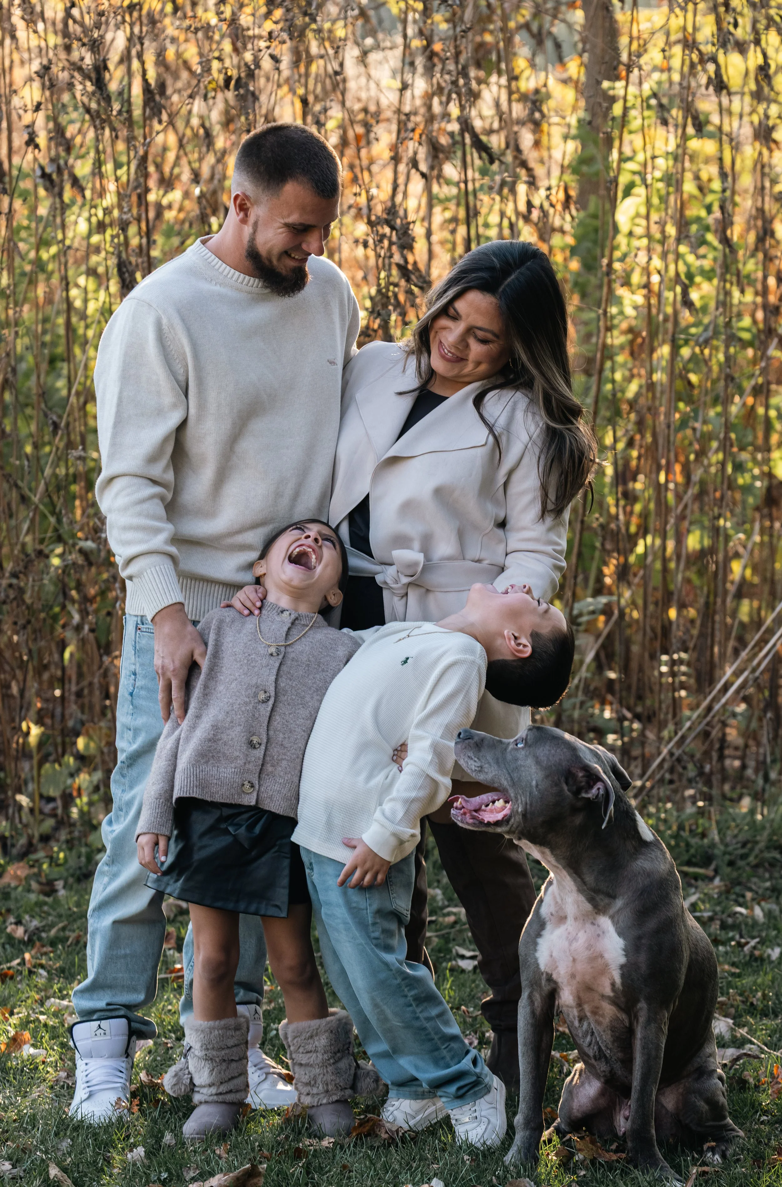 A smiling family of four, including two children and two adults, laughing and enjoying time outdoors in an autumn setting with fallen leaves and trees in the background, along with a large gray and white dog sitting on the grass.