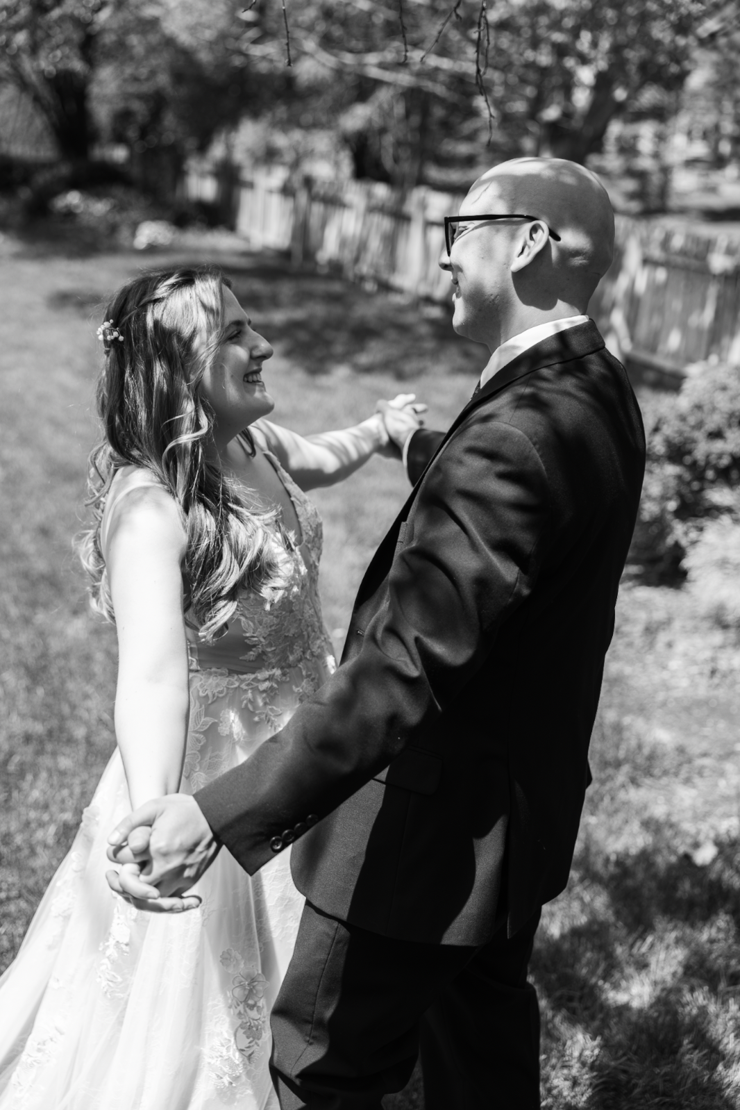 A black and white photo of a couple dancing outdoors. The woman is in a wedding dress and the man is in a suit, smiling at each other.