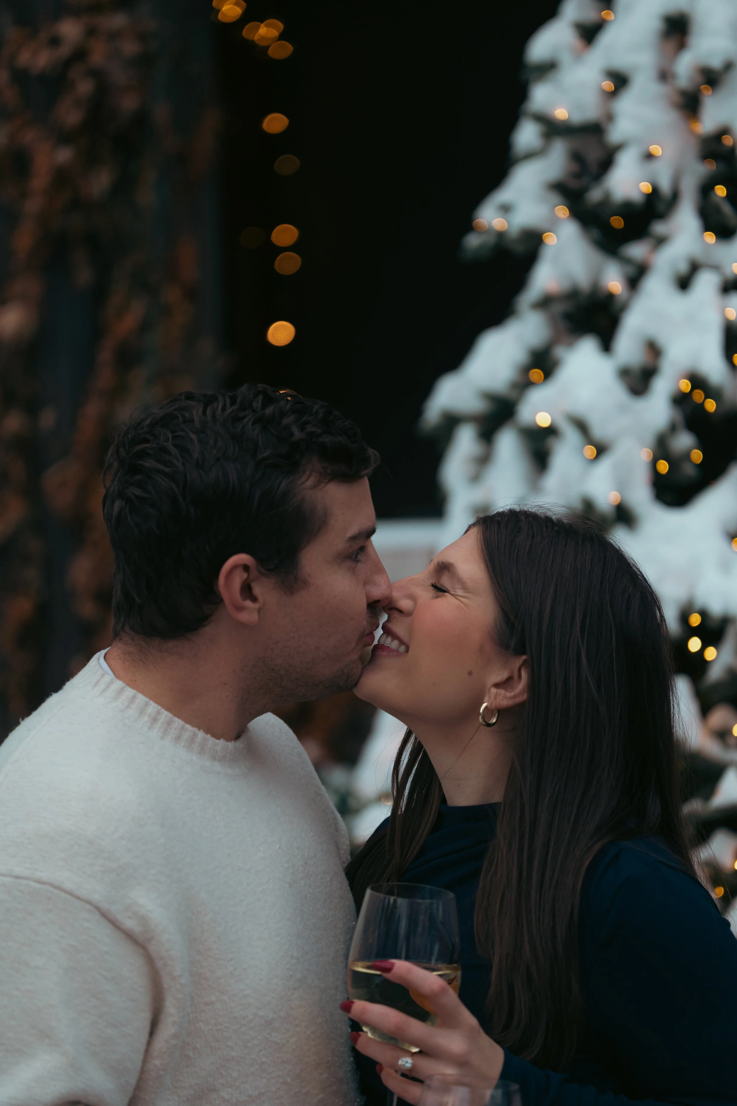 A couple stands close with their foreheads touching, smiling at each other during a festive gathering with Christmas lights and a decorated Christmas tree in the background. The woman holds a glass of white wine.