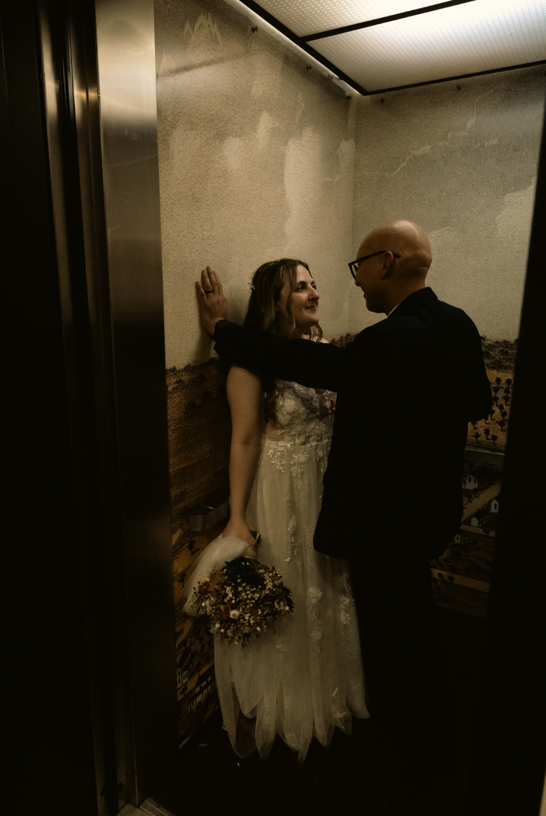 A bride and groom inside an elevator, sharing an intimate moment. The bride is holding a bouquet and wearing a wedding dress, while the groom is dressed in a dark suit. They are looking at each other affectionately.
