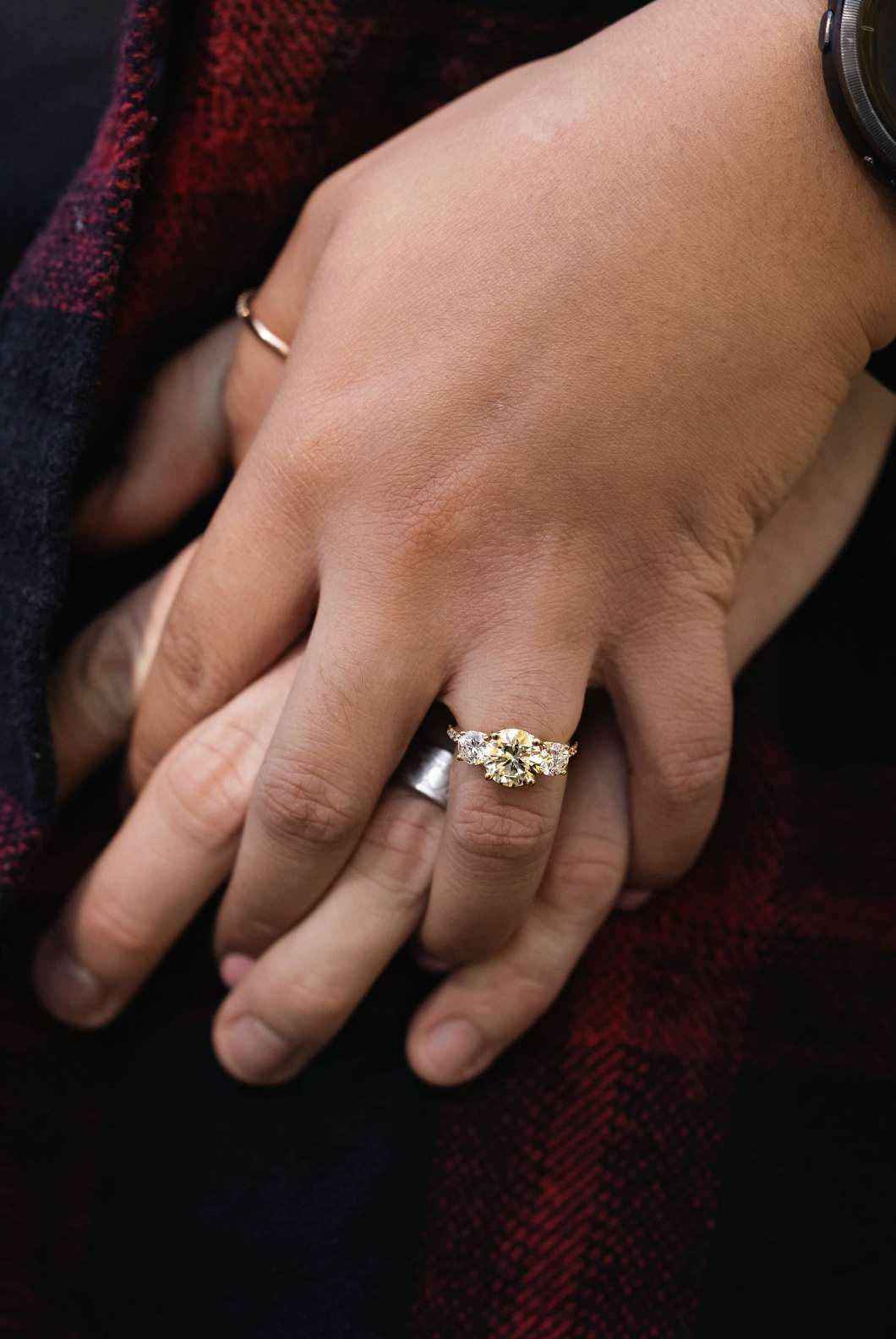 Close-up of a person's hand wearing a large diamond engagement ring, with a wedding band underneath, resting on another hand, all against a red and black plaid fabric background.