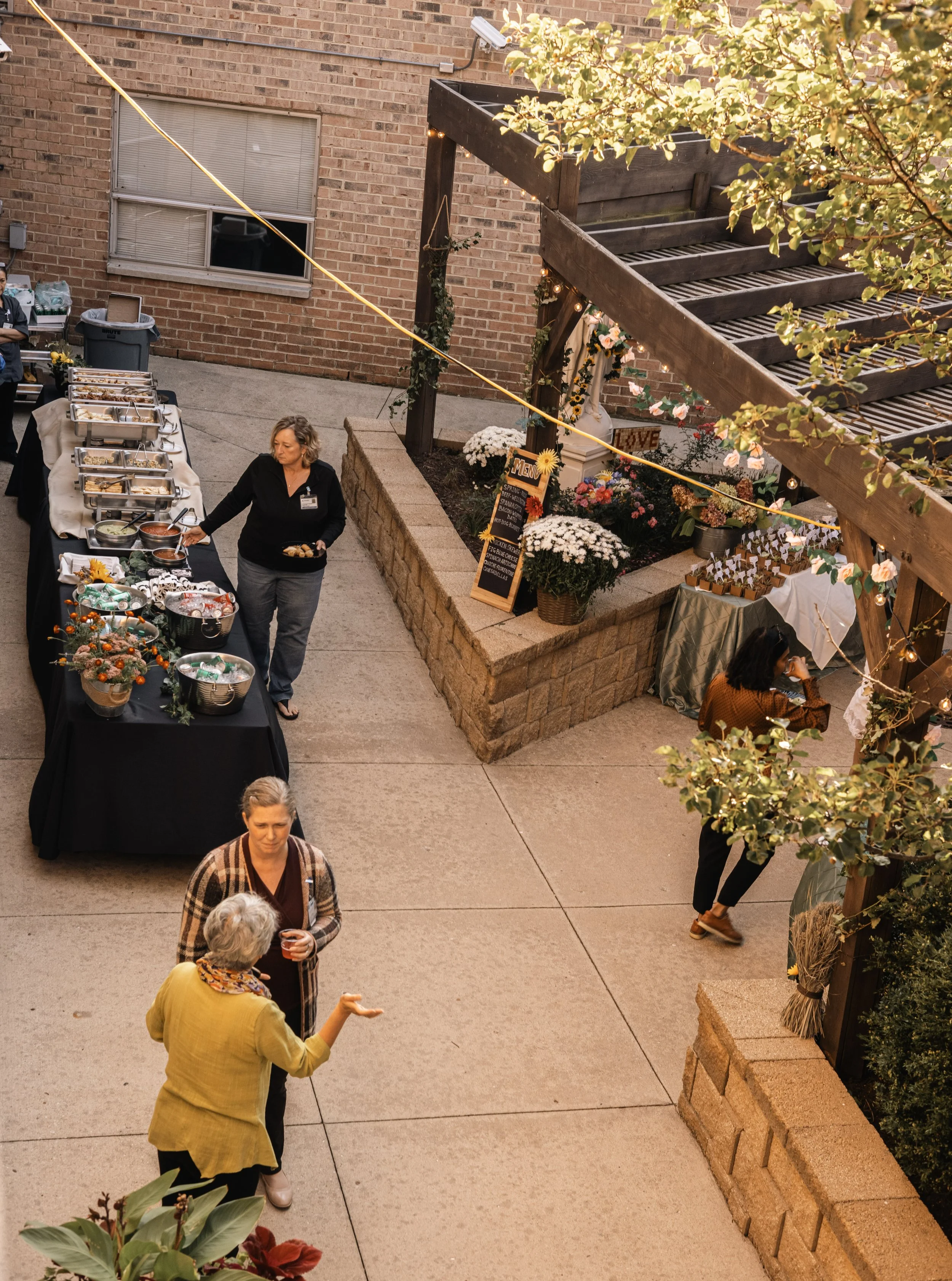 People attending an outdoor event, standing and conversing near tables with food, flowers, and decorations, with warm lighting and a brick building in the background.