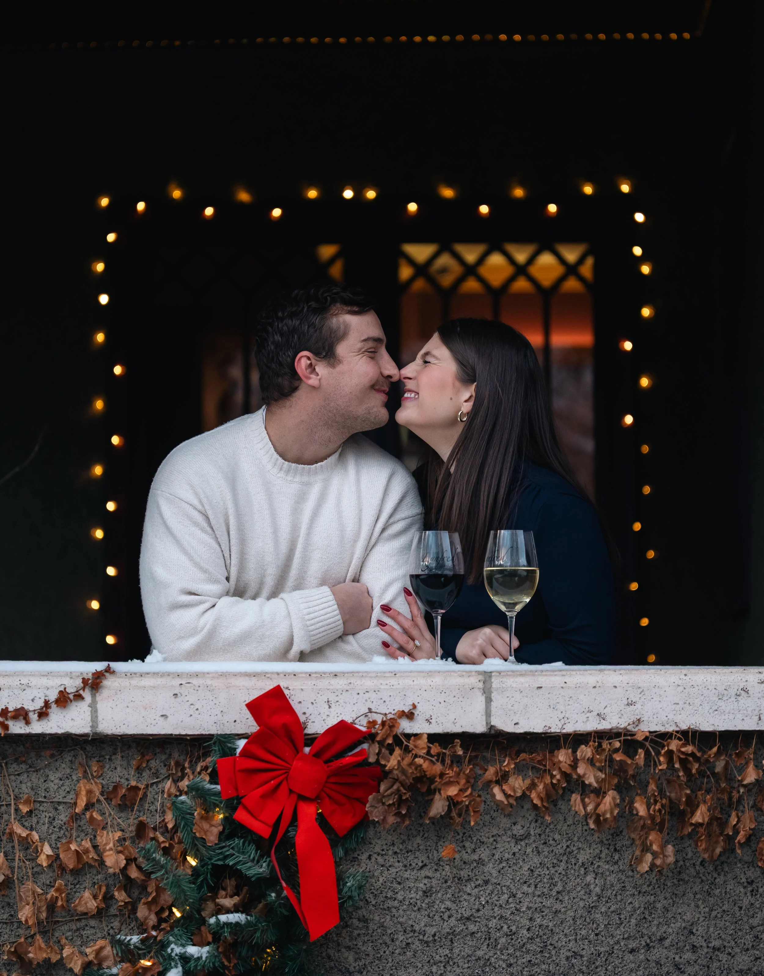 A couple smiling and leaning close to each other with noses touching, holding wine glasses in a cozy, decorated environment with holiday lights and wreaths.