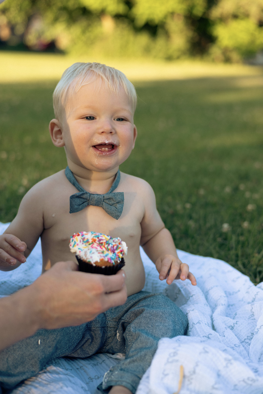 A young boy with blond hair and a grey bowtie, sitting on a blanket outdoors, smiling as he receives a cupcake with sprinkles.