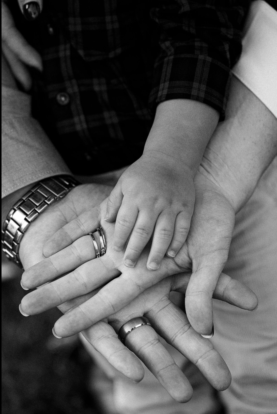 A black and white photo of two people, an adult and a child, holding hands. The adult's hand has a watch and wedding band, while the child's hand rests on top of it, all fingers intertwined.