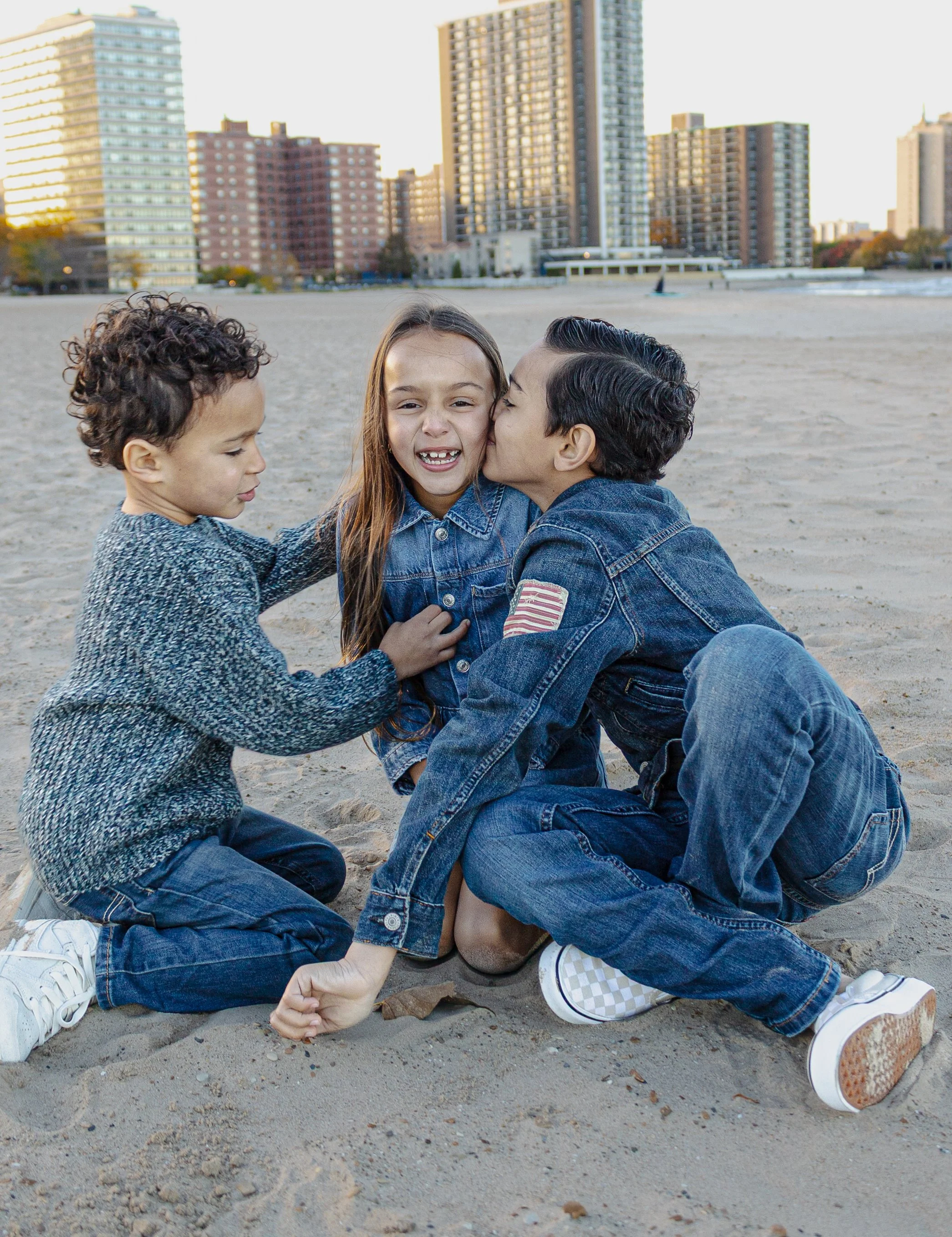 Three children sitting on a sandy beach with tall buildings in the background. One girl is being kissed on the cheek by a boy, and another boy is sitting nearby.