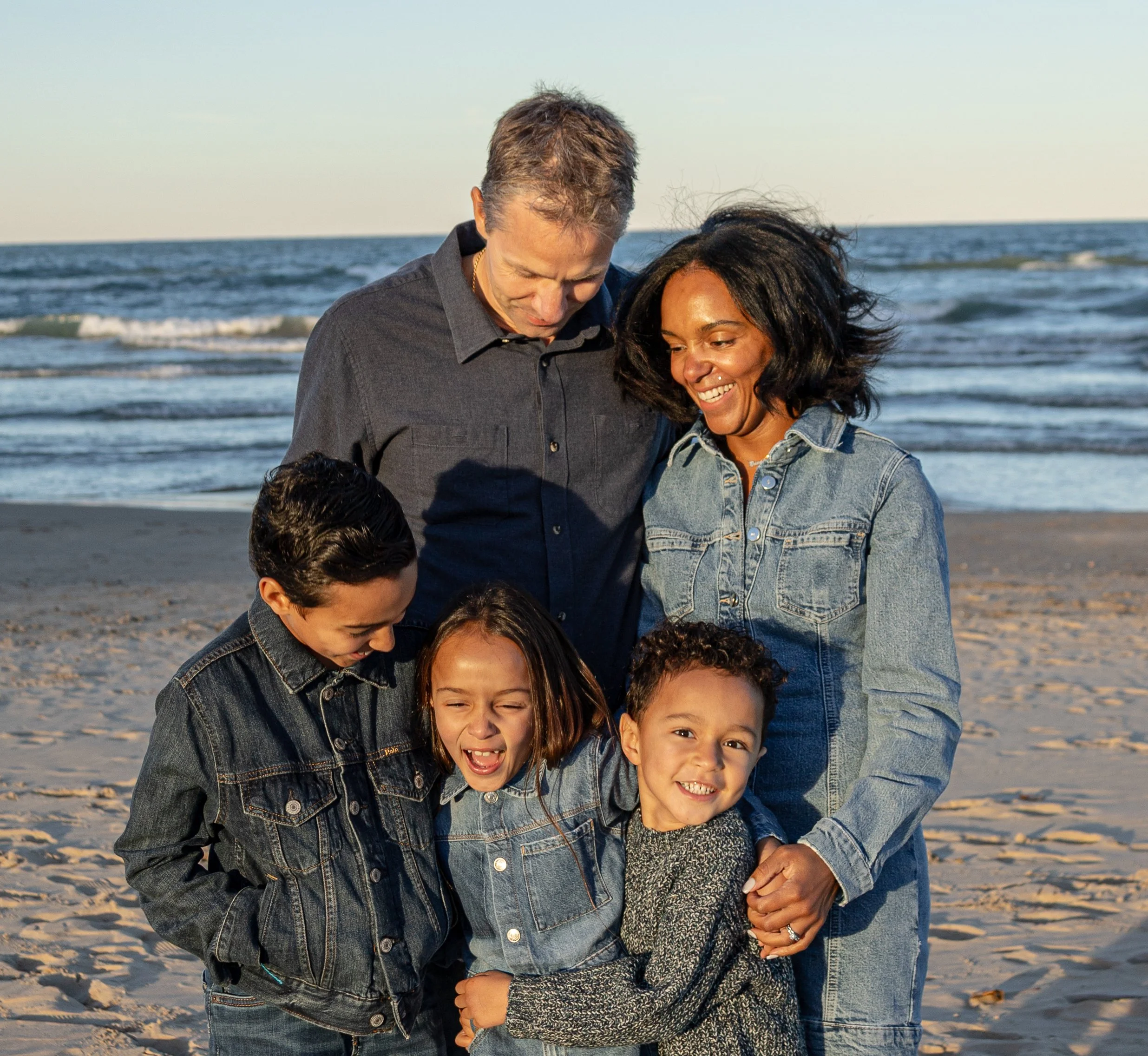 A group of children and two adults smiling and hugging on a beach near the ocean with gentle waves.