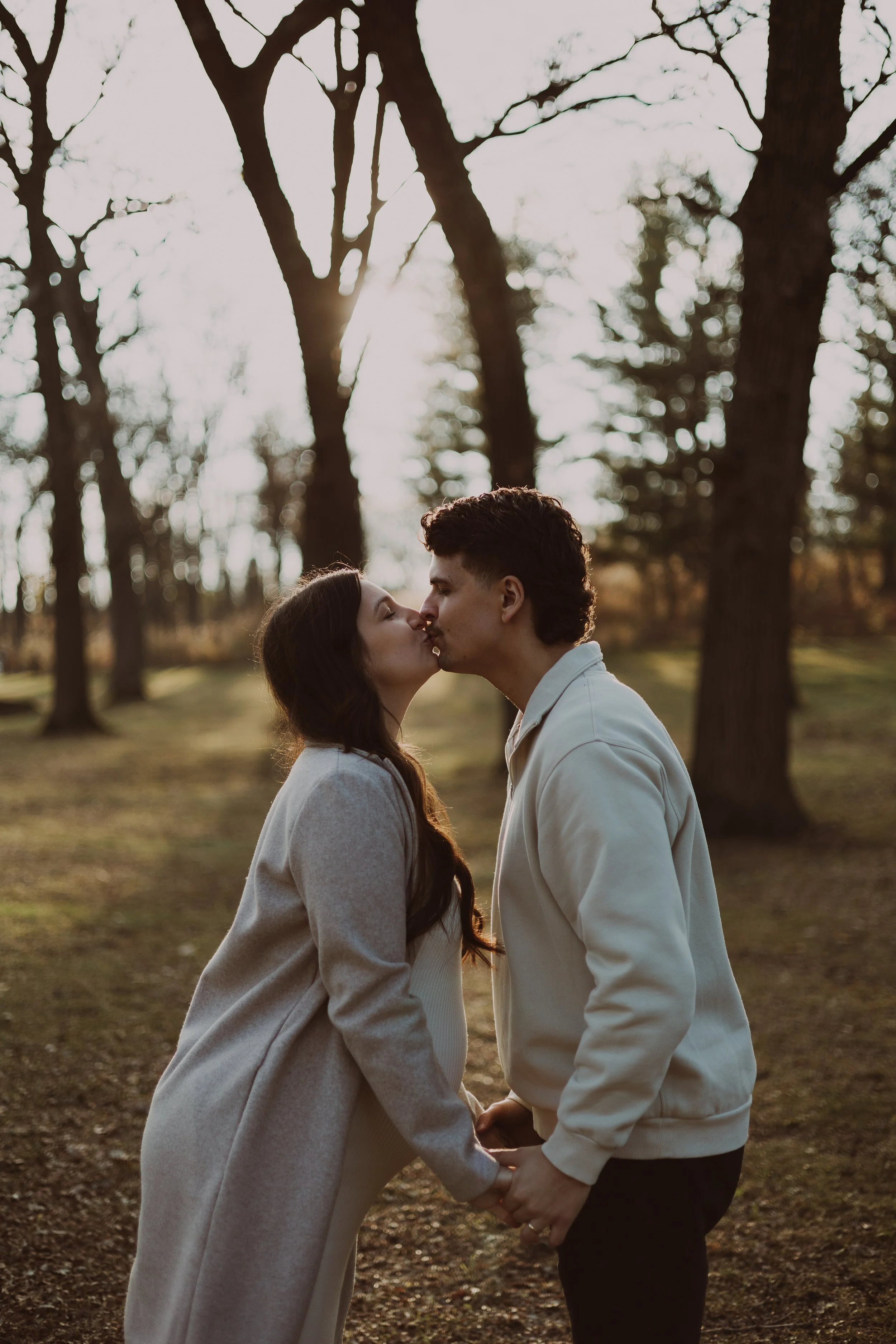A young couple stands closely together outdoors during sunset, holding hands and about to kiss, surrounded by tall trees in a park or forest setting.