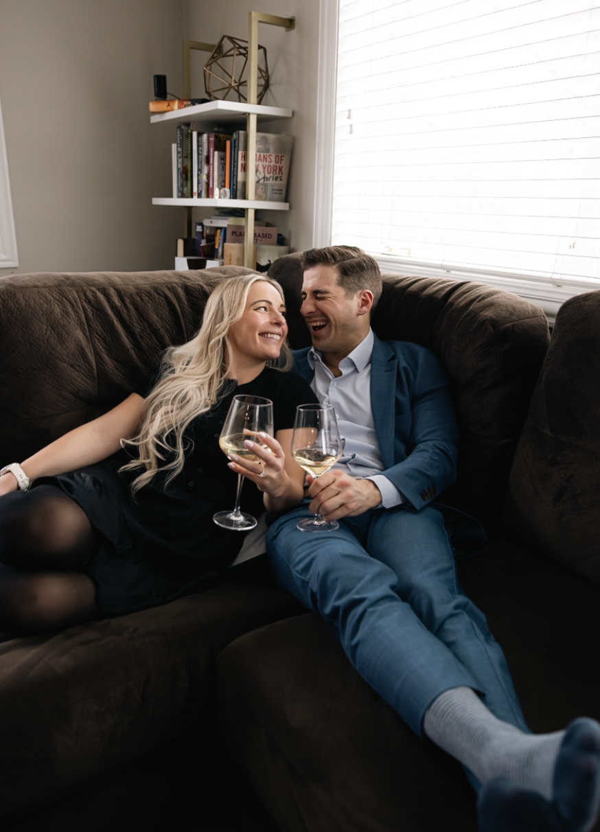 A couple sitting on a brown couch, smiling and laughing while holding glasses of white wine, enjoying each other's company in a cozy living room.