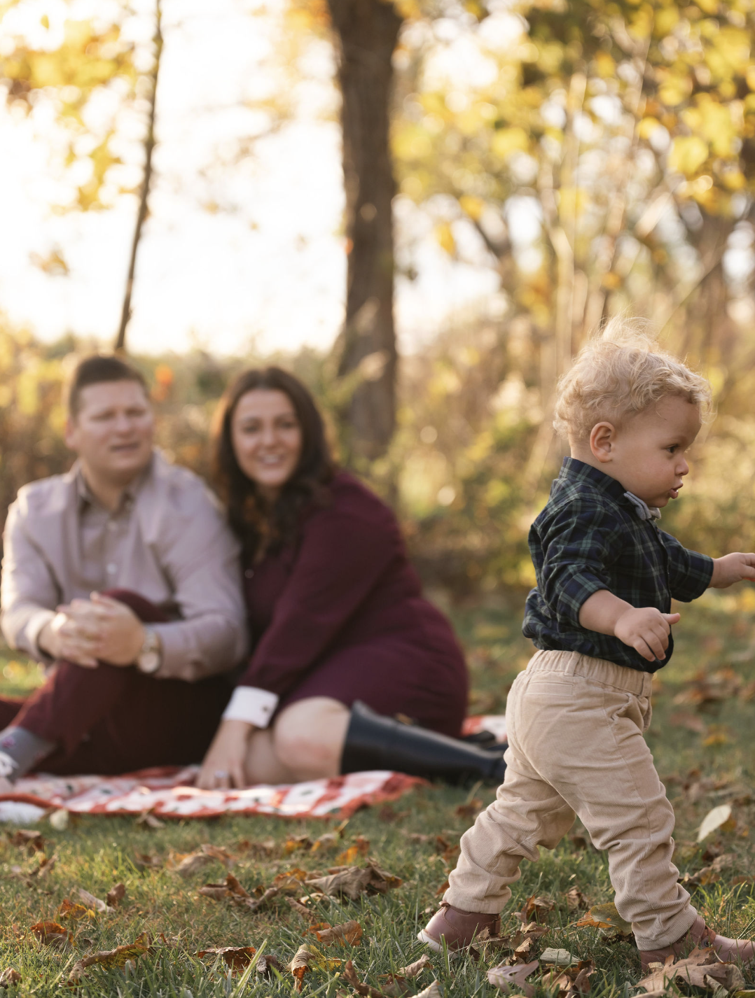 A young boy walking in a park with fallen leaves, with a man and woman sitting on a blanket in the background during autumn.