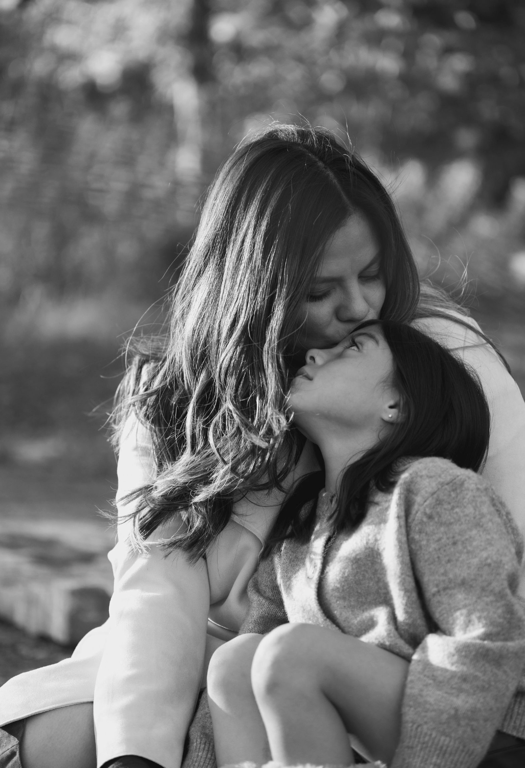 Black and white photograph of a mother and daughter sharing an affectionate moment outdoors, with the mother kissing the daughter's forehead as they sit close together.