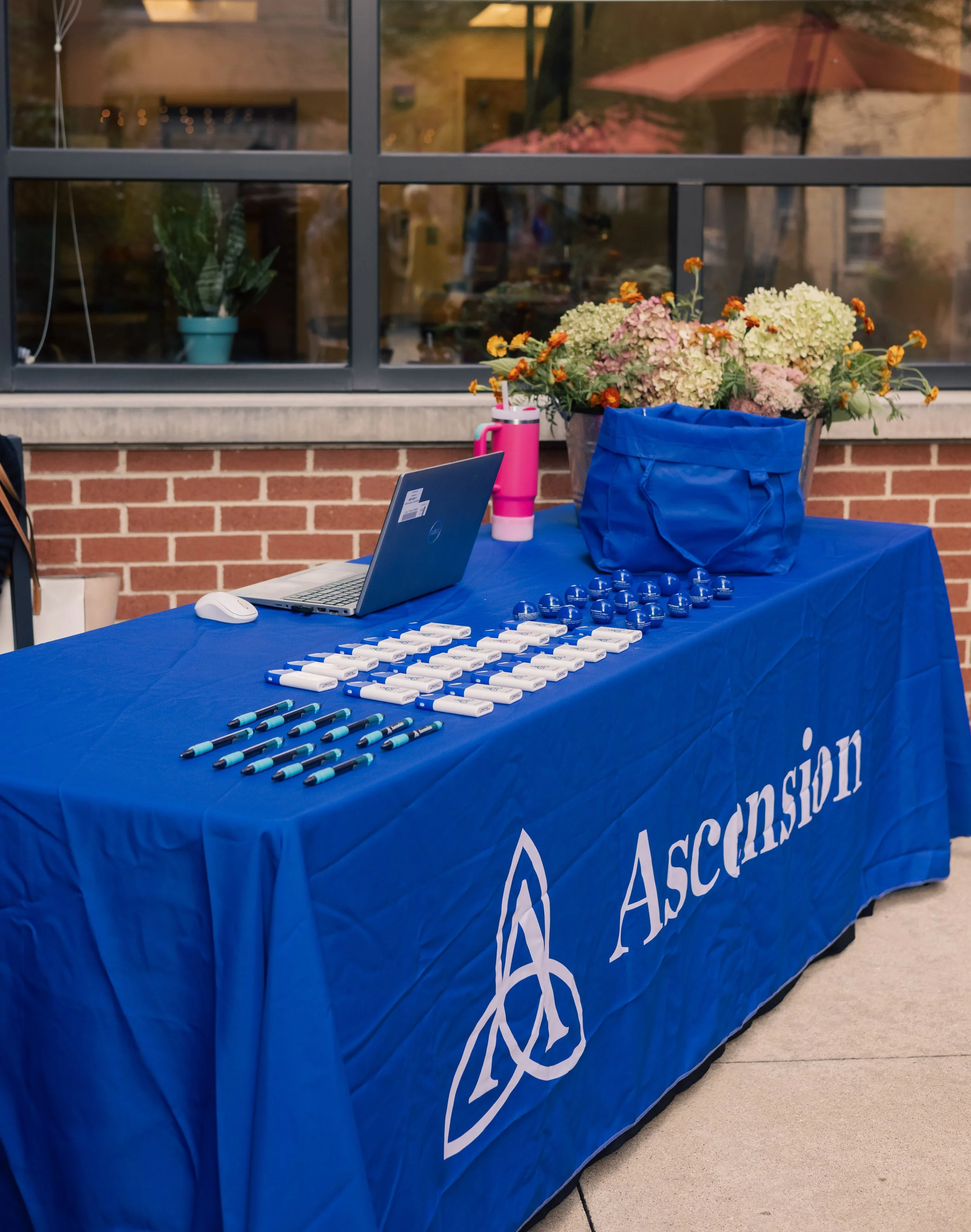 A blue tablecloth with the organizations logo, covered with whiteboards, blue markers, and aloe water bottles, placed in front of a window with floral arrangements visible indoors and outdoors.